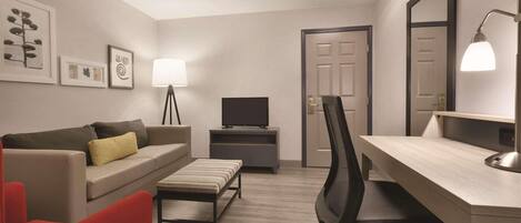A modern hotel room with a sofa, a red chair, a desk with a computer, a television, and a mirror.