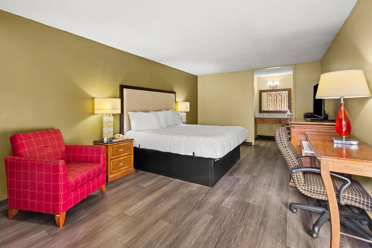 A hotel room with a bed, a red armchair, a desk with a chair, and a wooden dresser.
