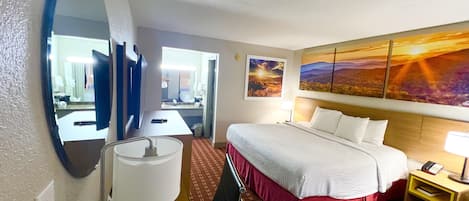 A hotel room with a bed, a desk, a chair, a mirror, a lamp, a nightstand, and a painting on the wall.