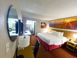 Habitación de hotel con cama, escritorio, silla, espejo, lámpara, mesita de noche y un cuadro en la pared.