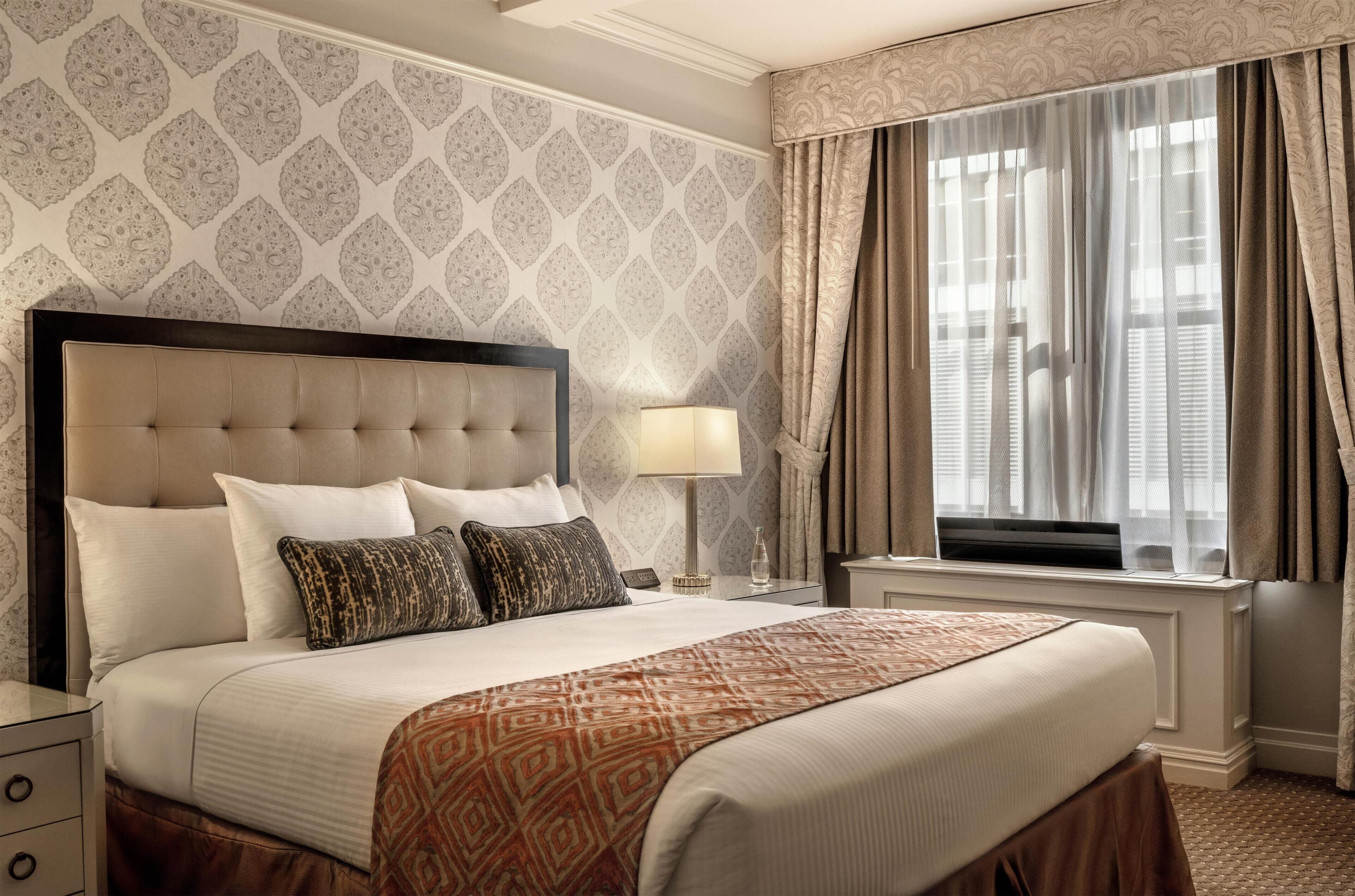 Quarto de hotel com uma cama grande, uma mesa de cabeceira com abajur, uma televisão e uma janela com cortinas.