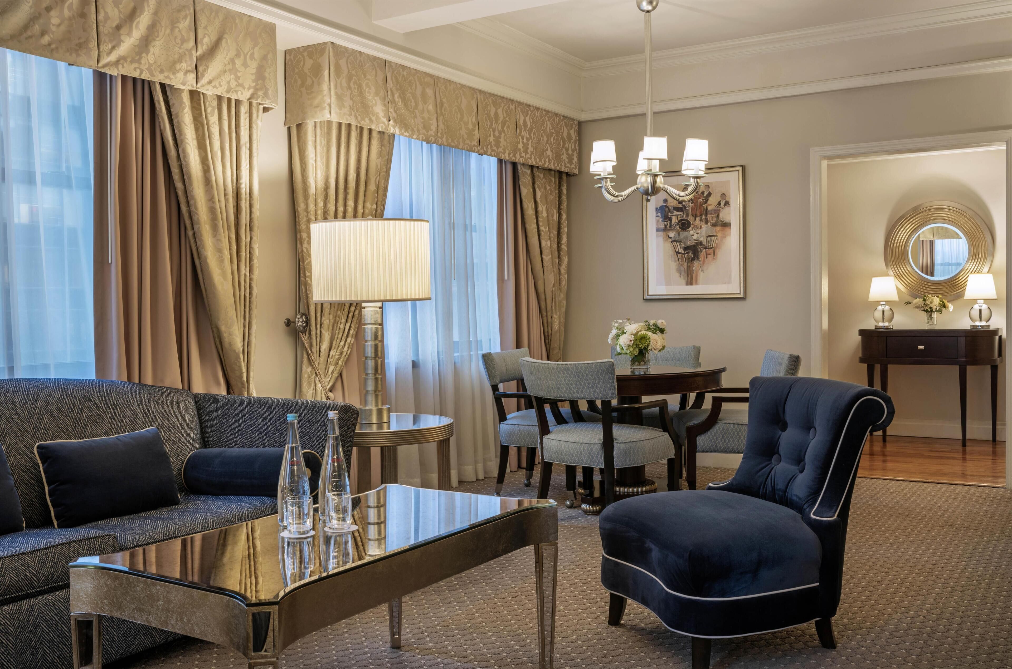 A hotel room with a sofa, armchair, dining table, chairs, a lamp, a mirror, and a chandelier.