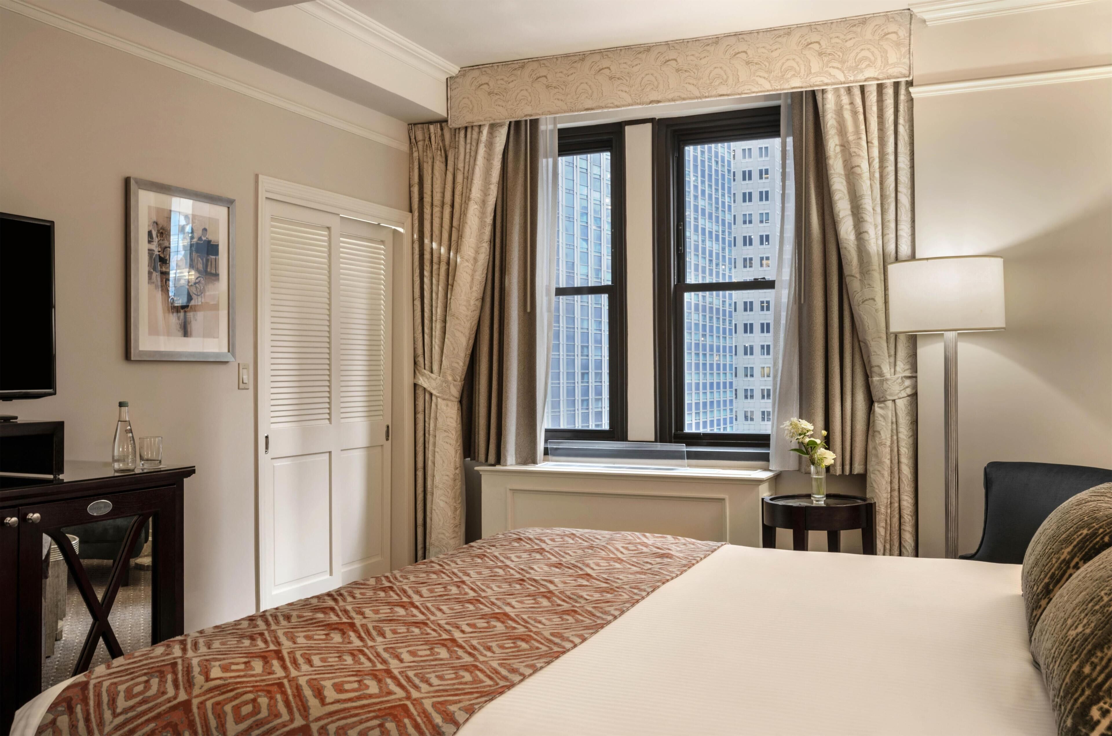 Una habitación de hotel con cama, televisor, ventana con cortinas, lámpara y un florero con flores.