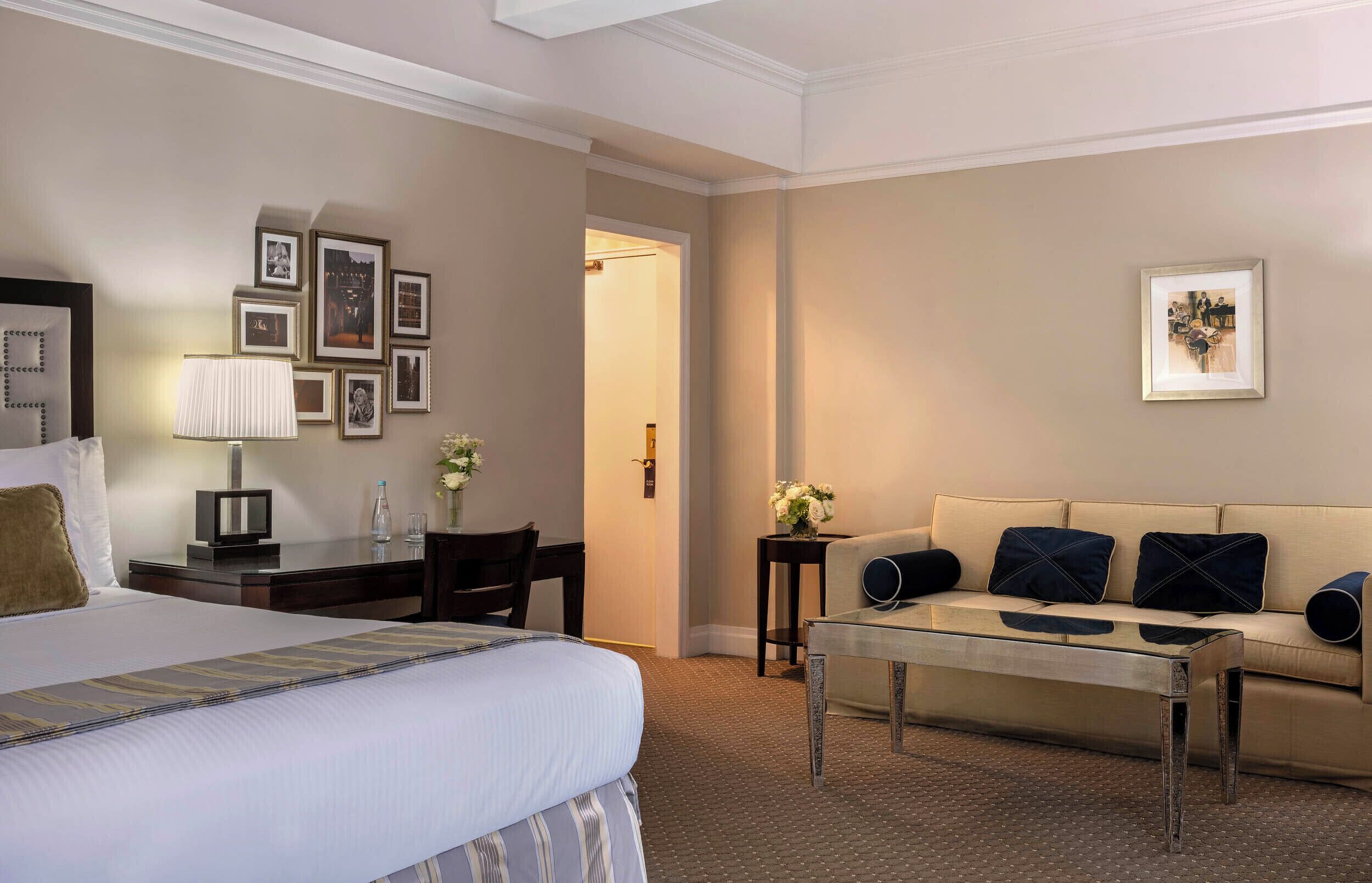 A hotel room with a bed, a sofa, a coffee table, a desk with a lamp, and framed pictures on the walls.