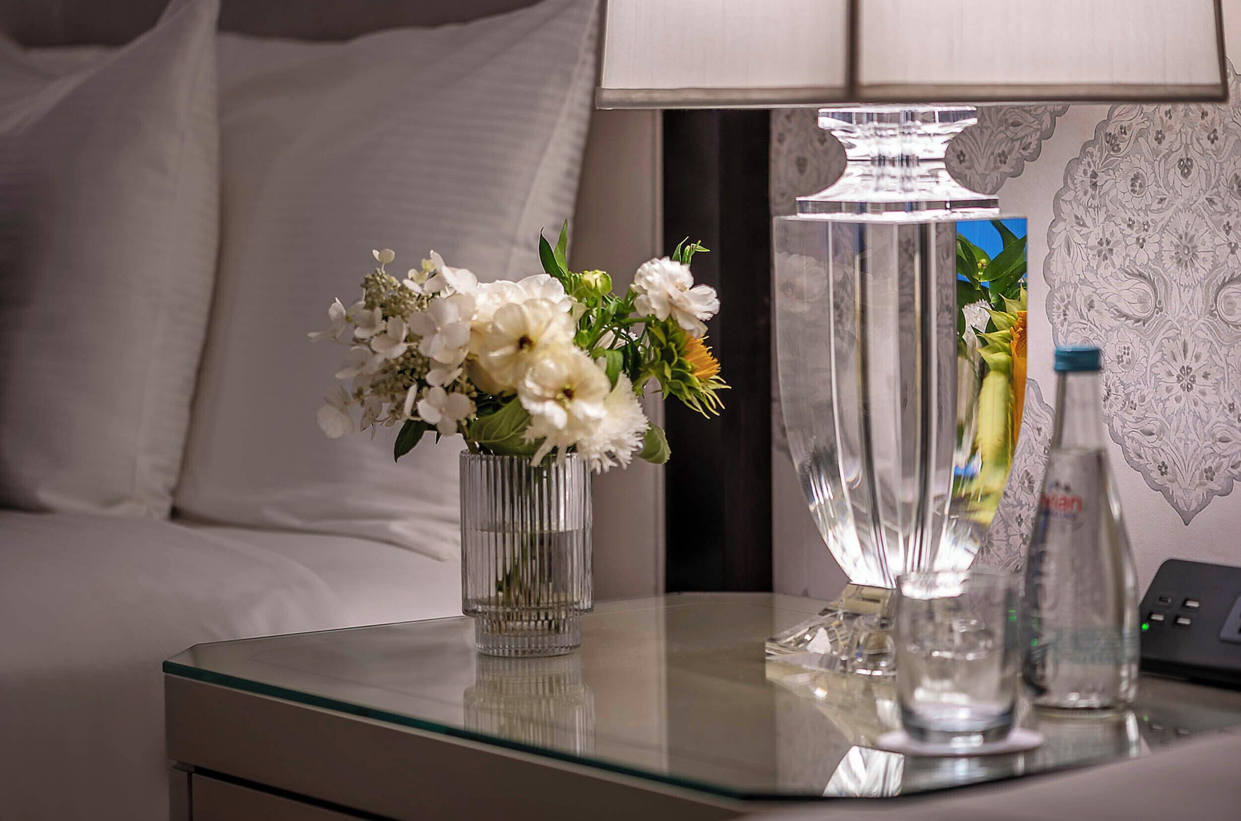 Una habitació d'hotel amb un llit, una taula de vidre amb un ram de flors, una làmpada i una ampolla d'aigua.