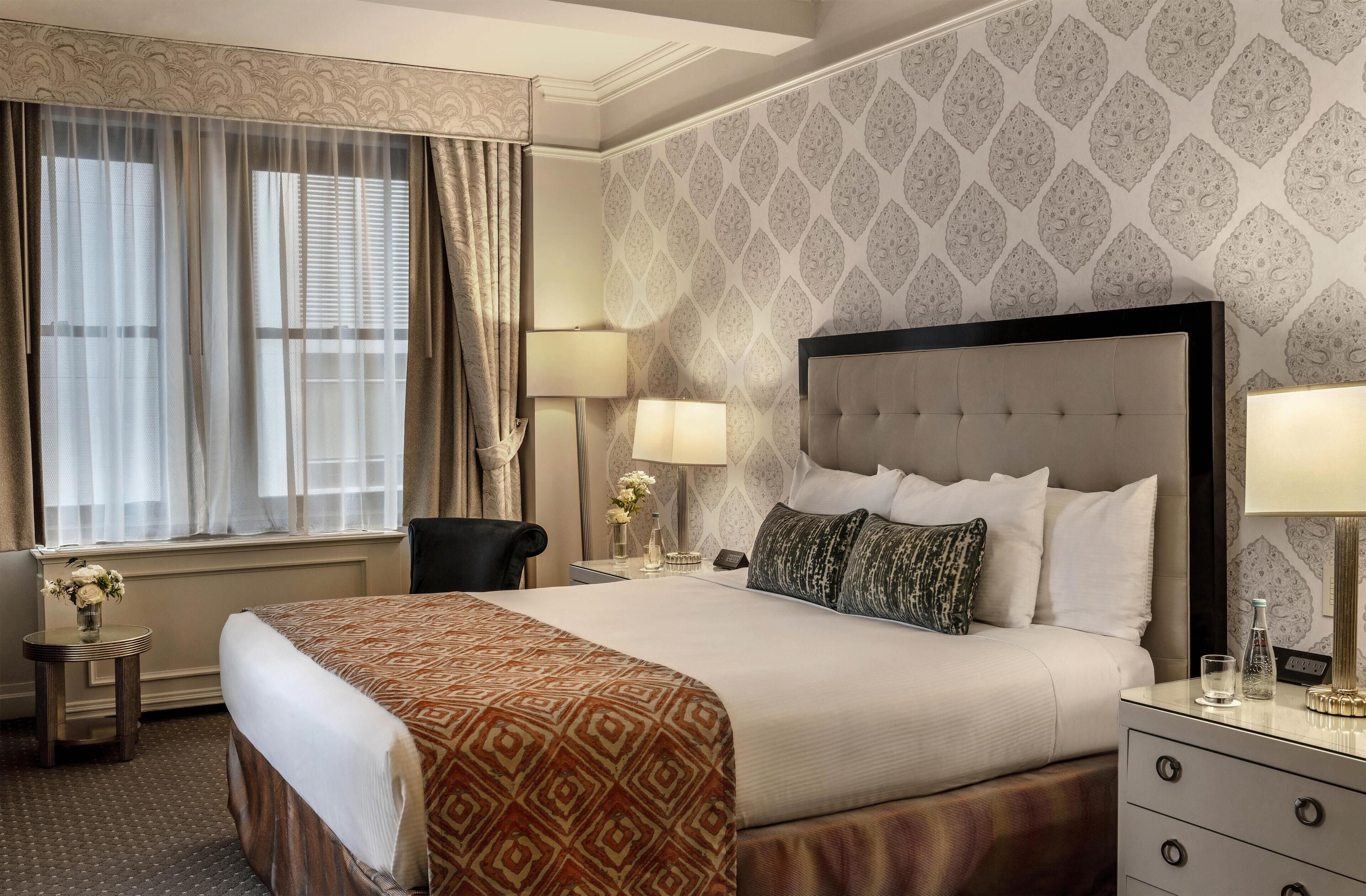 Habitación de hotel con una cama grande, dos mesitas de noche, una silla y una ventana con cortinas.