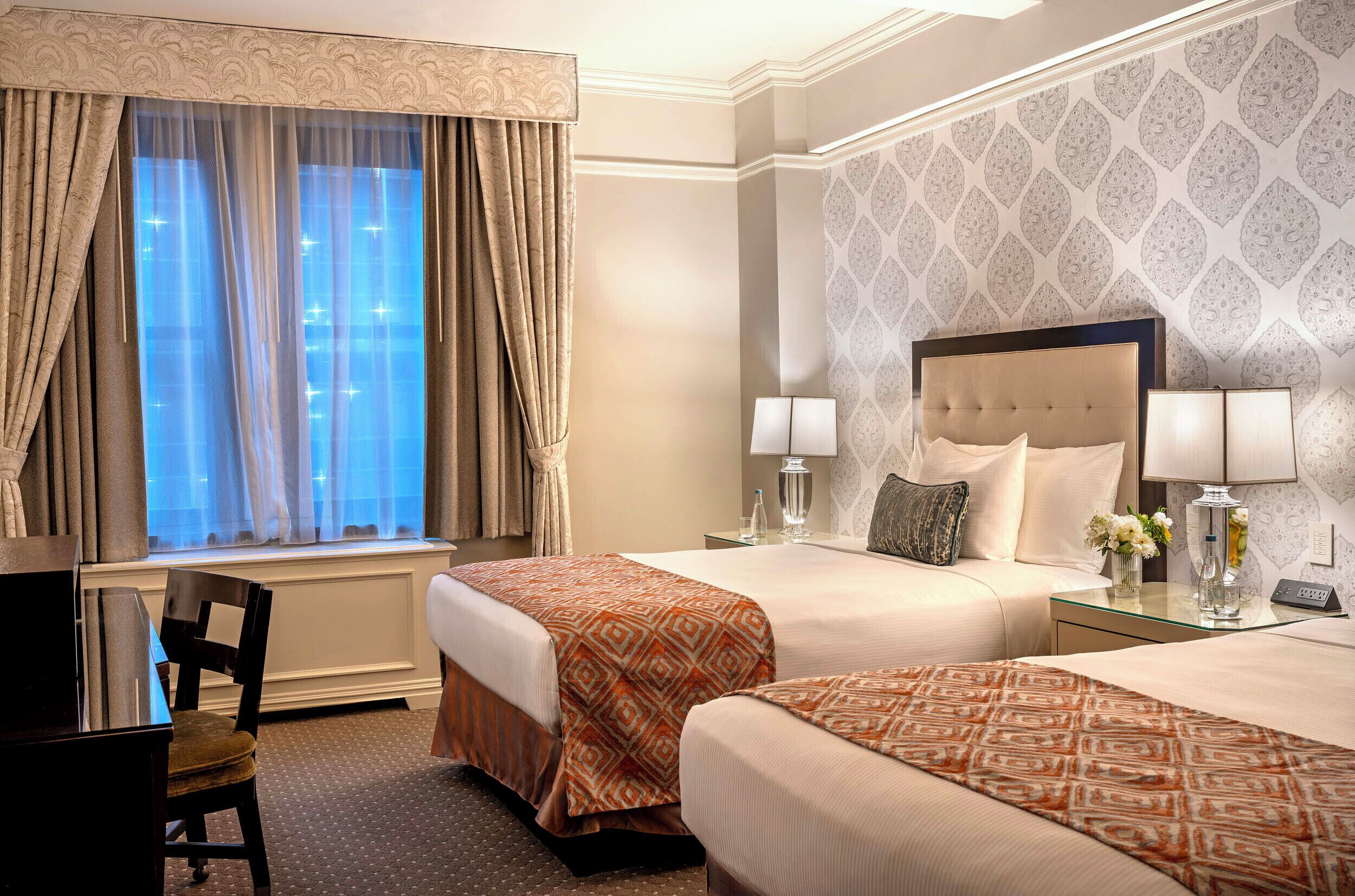 A hotel room with two beds, a desk, a chair, and a window with curtains.