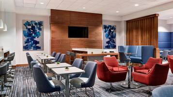 Een moderne hotel lobby met een lange receptiebalie, blauwe en rode zitplaatsen, en een groot televisiescherm.