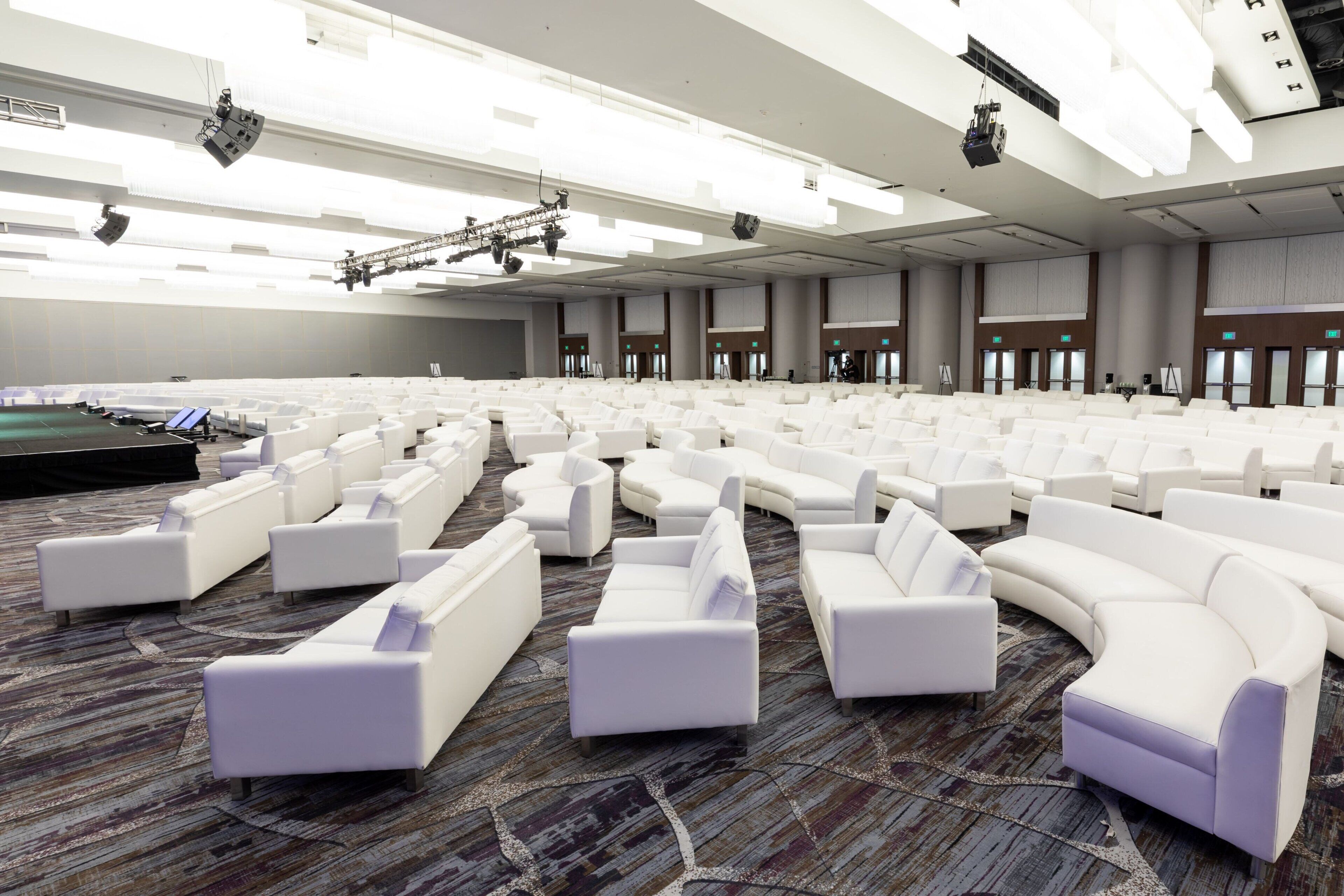 A large conference room with white sofas arranged in rows, a stage with lighting equipment, and a carpeted floor.