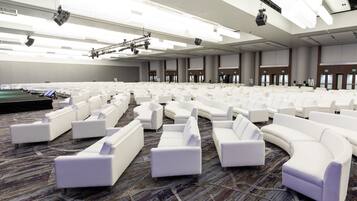 A large conference room with white sofas arranged in rows, a stage with lighting equipment, and a carpeted floor.