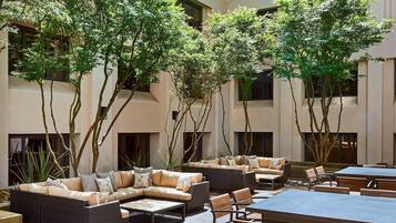 An outdoor seating area with sofas and tables, surrounded by trees and a building facade.