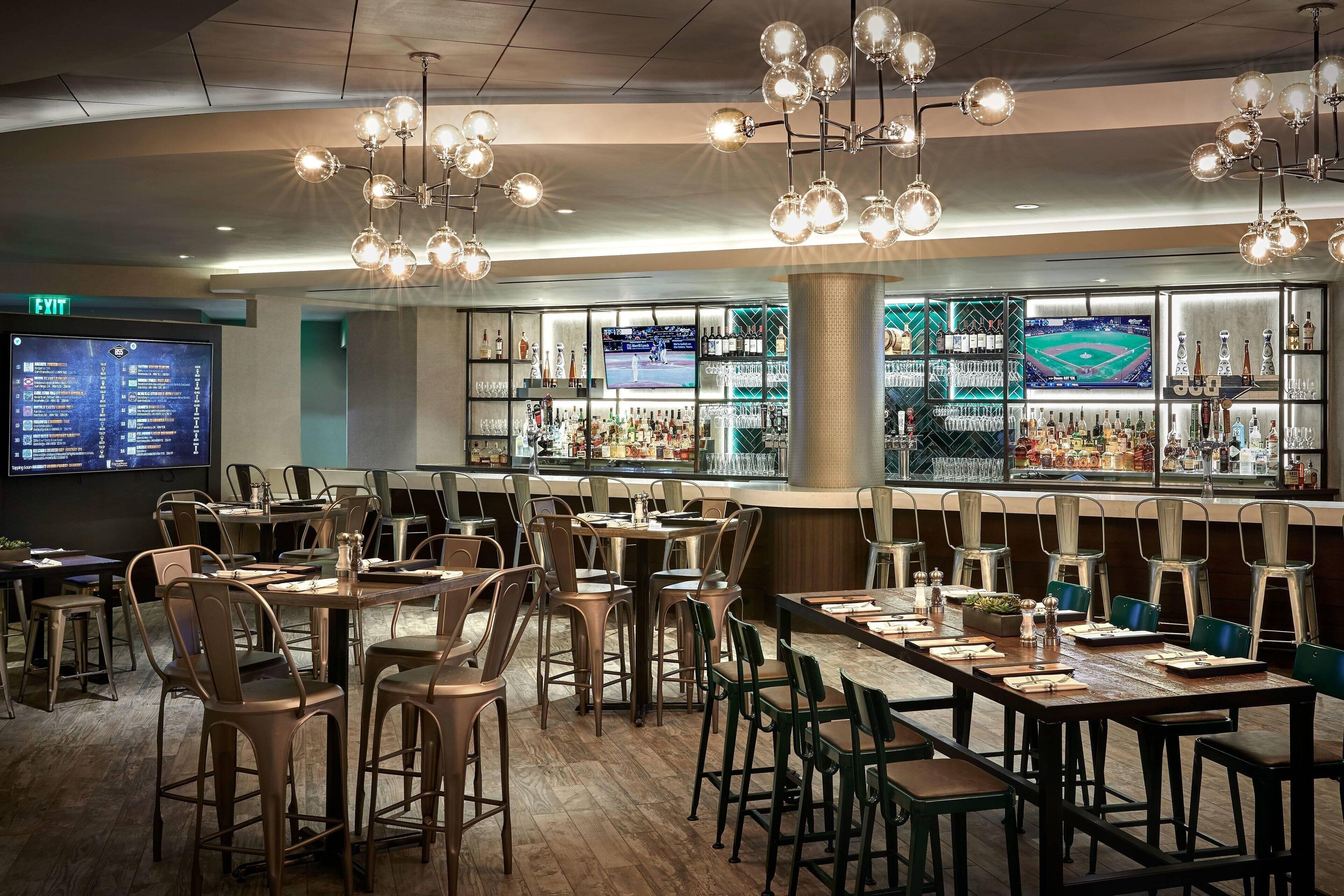 A restaurant with multiple tables and chairs, a bar area with shelves of bottles, and a large screen displaying a sports event.