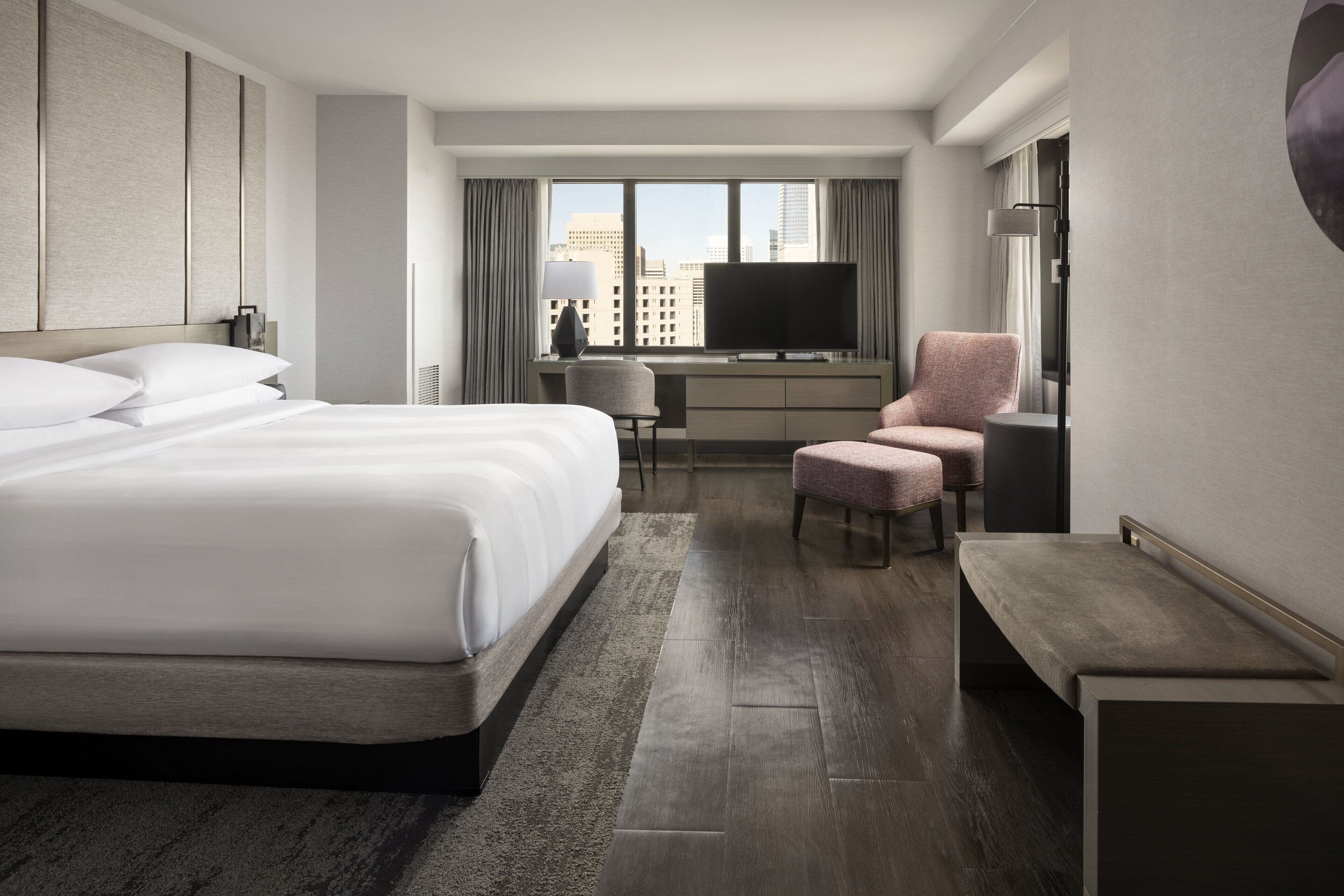 A modern hotel room with a large bed, a desk, a television, and a city view.