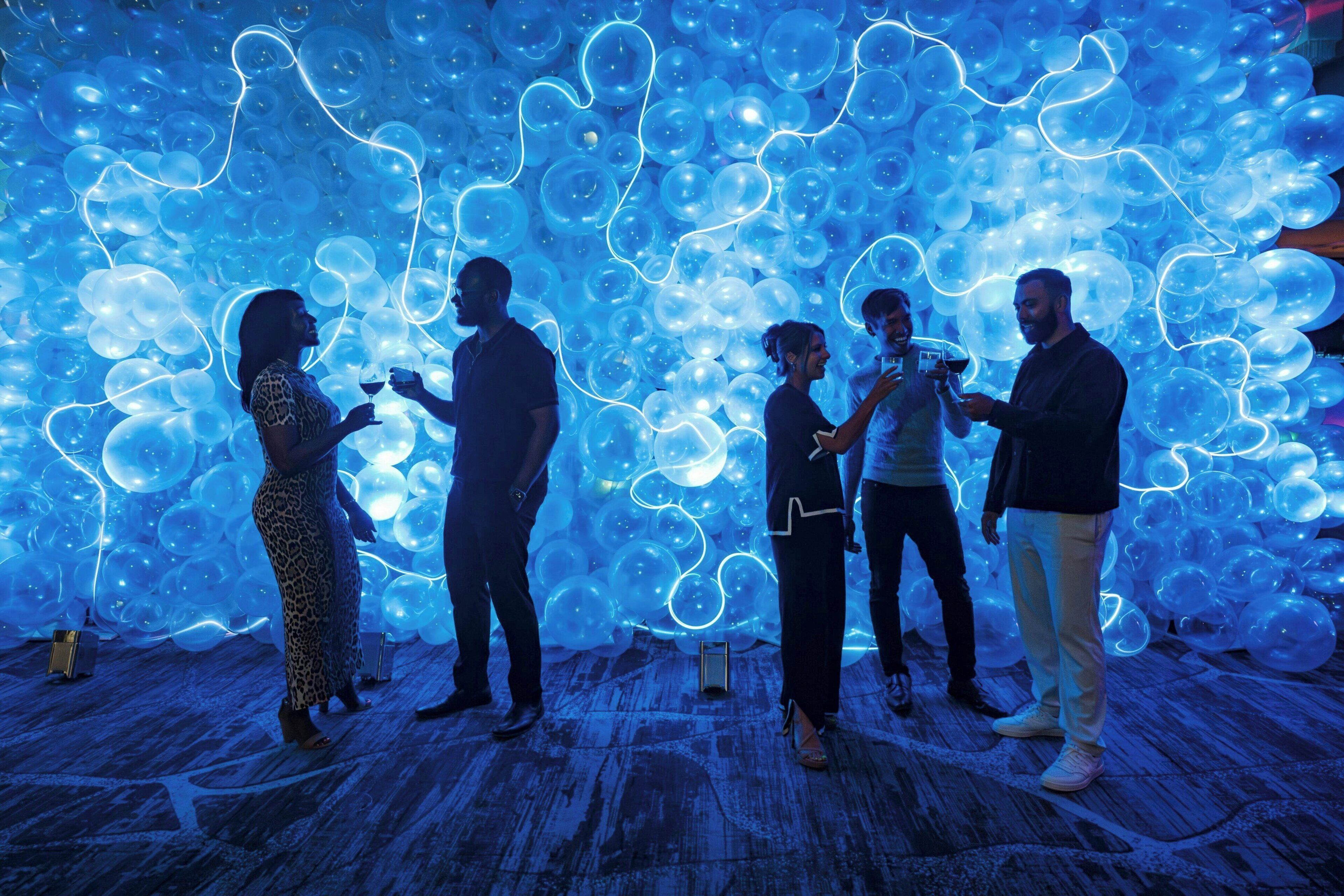 A room with a wooden floor, a large wall of blue balloons, and a few people standing.