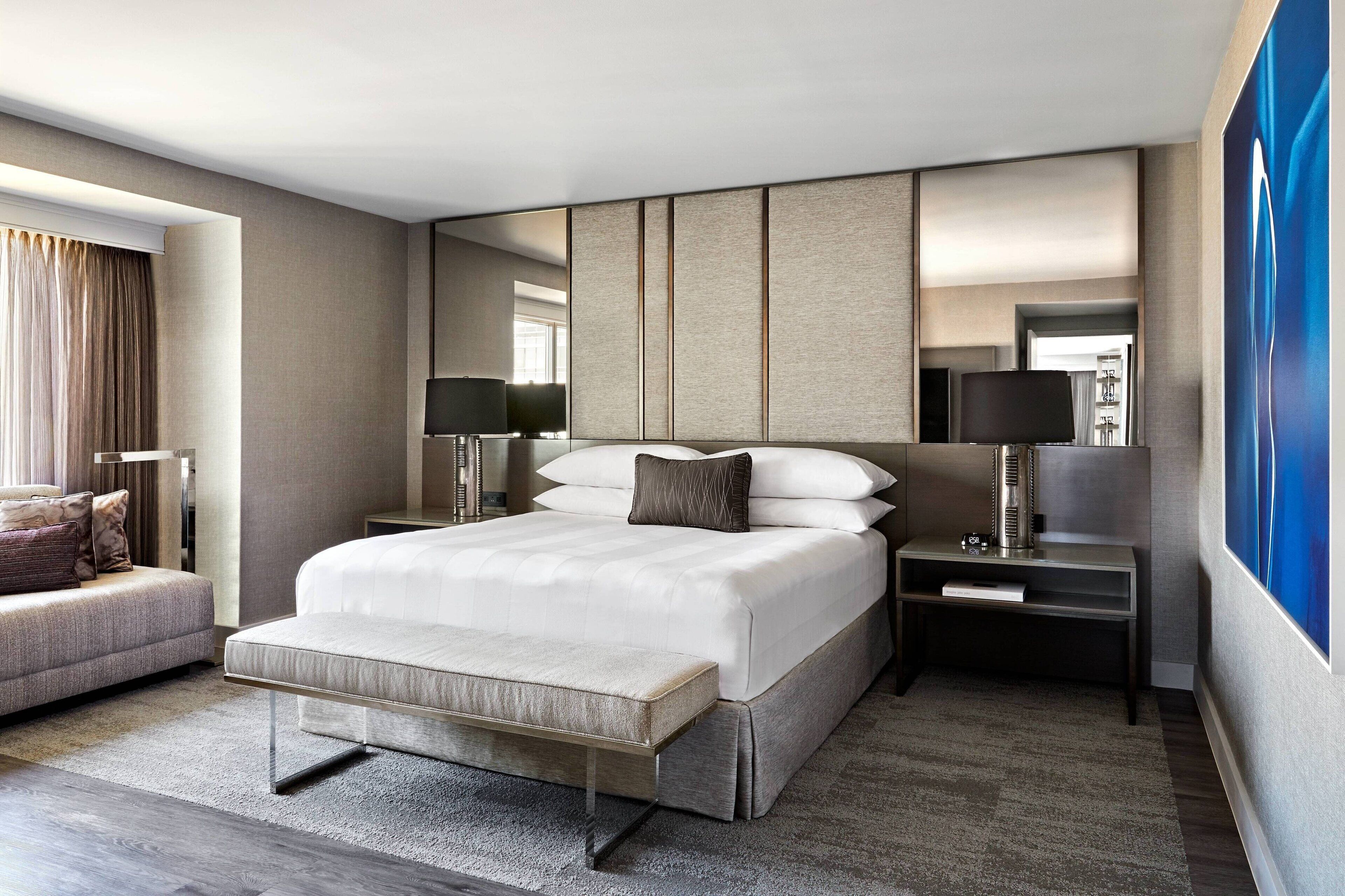 A modern hotel room with a large bed, bedside tables, a sofa, and a large window with curtains.
