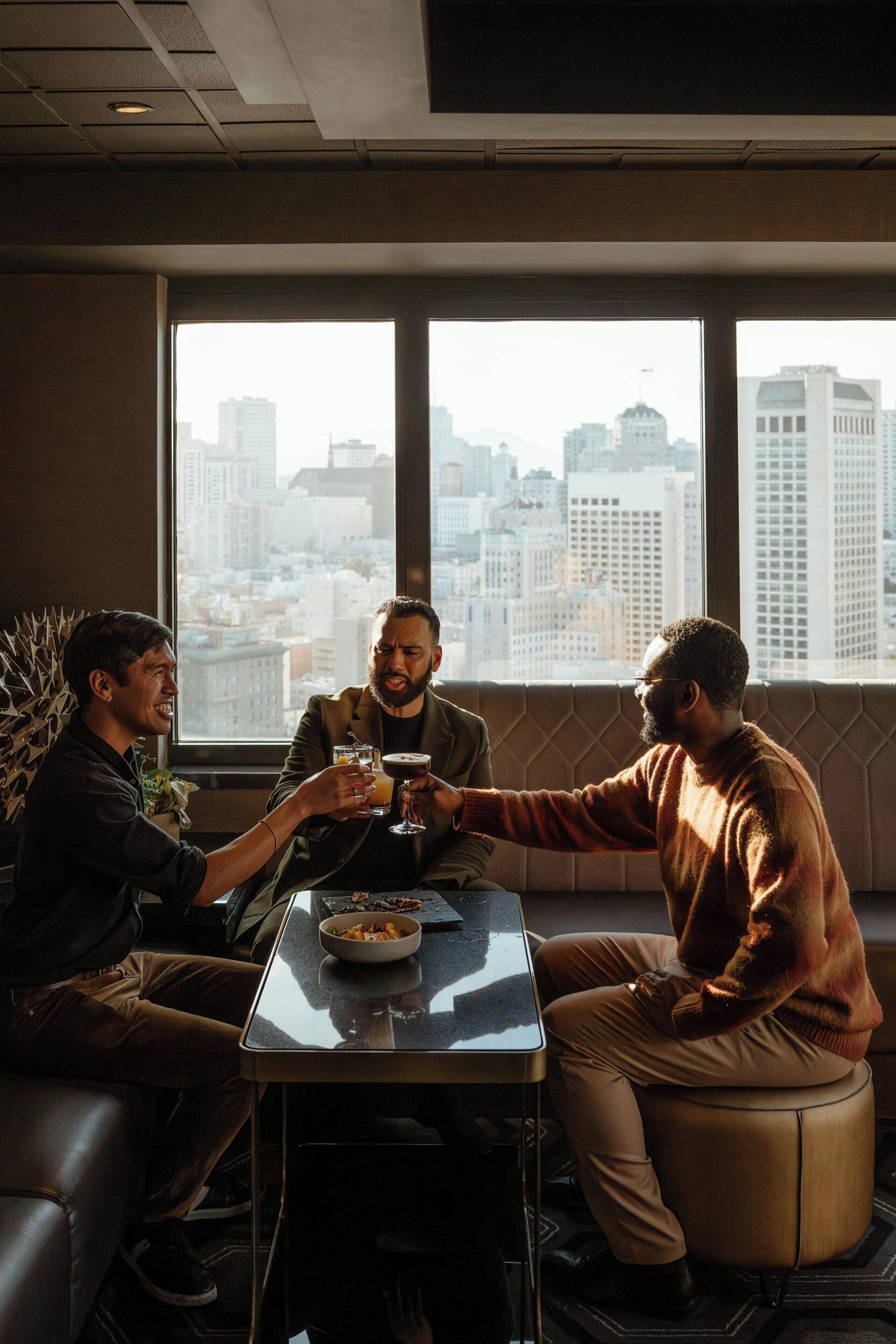 Three people toasting at a table with a city view through large windows.