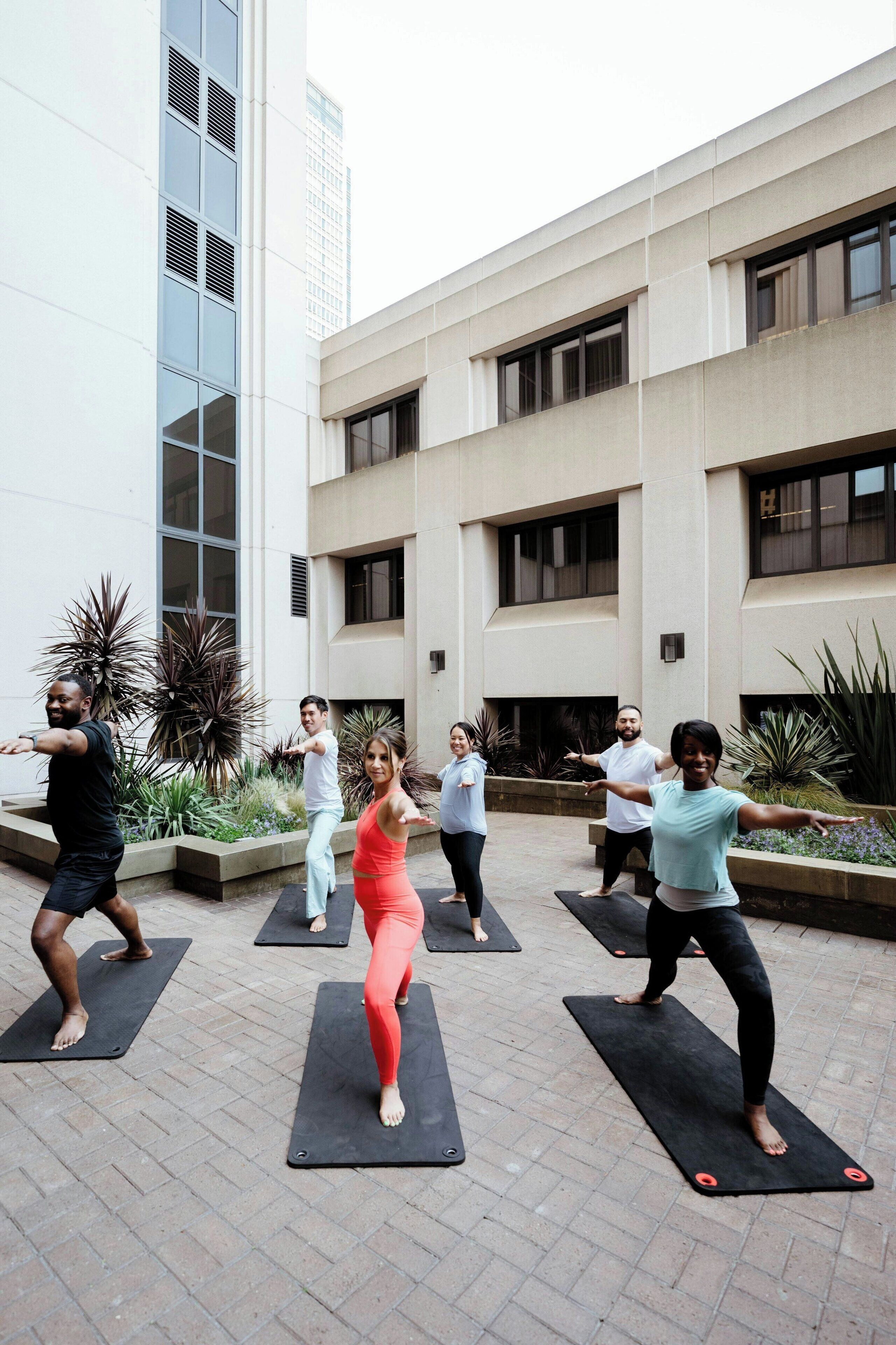 A group of people performing a yoga exercise outdoors in front of a building with large windows and a tiled floor.