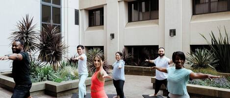A group of people performing a yoga exercise outdoors in front of a building with large windows and a tiled floor.