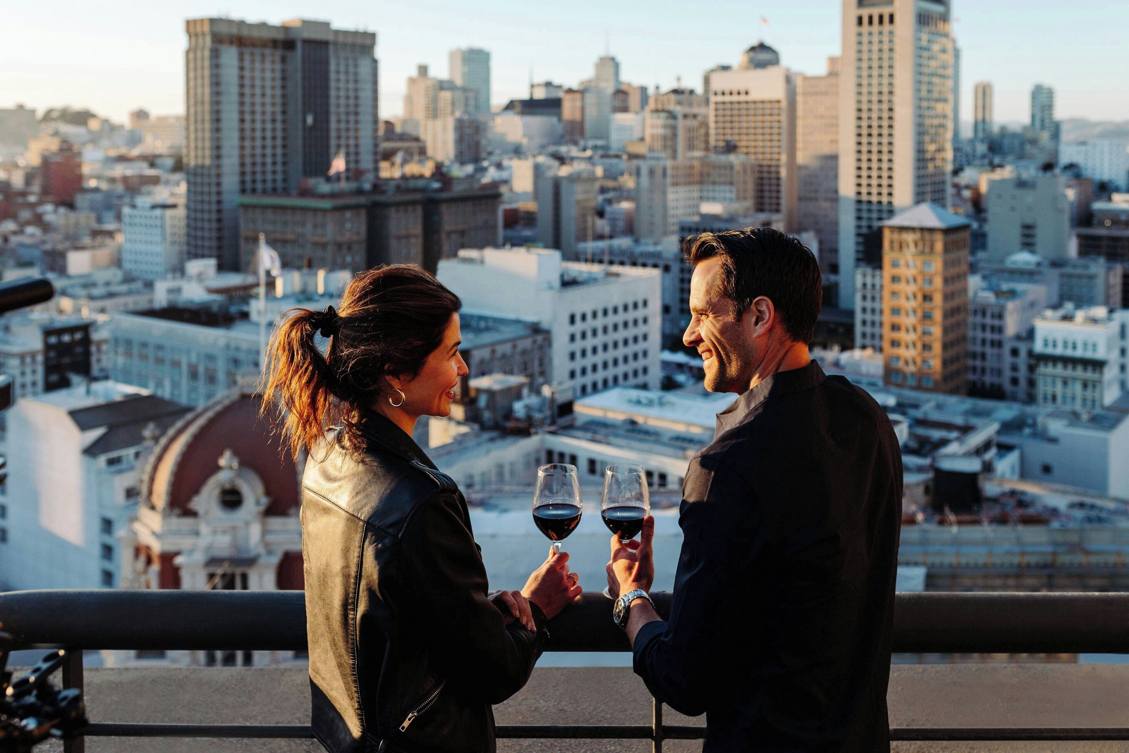 A couple toasting with wine glasses on a balcony overlooking a cityscape with tall buildings.