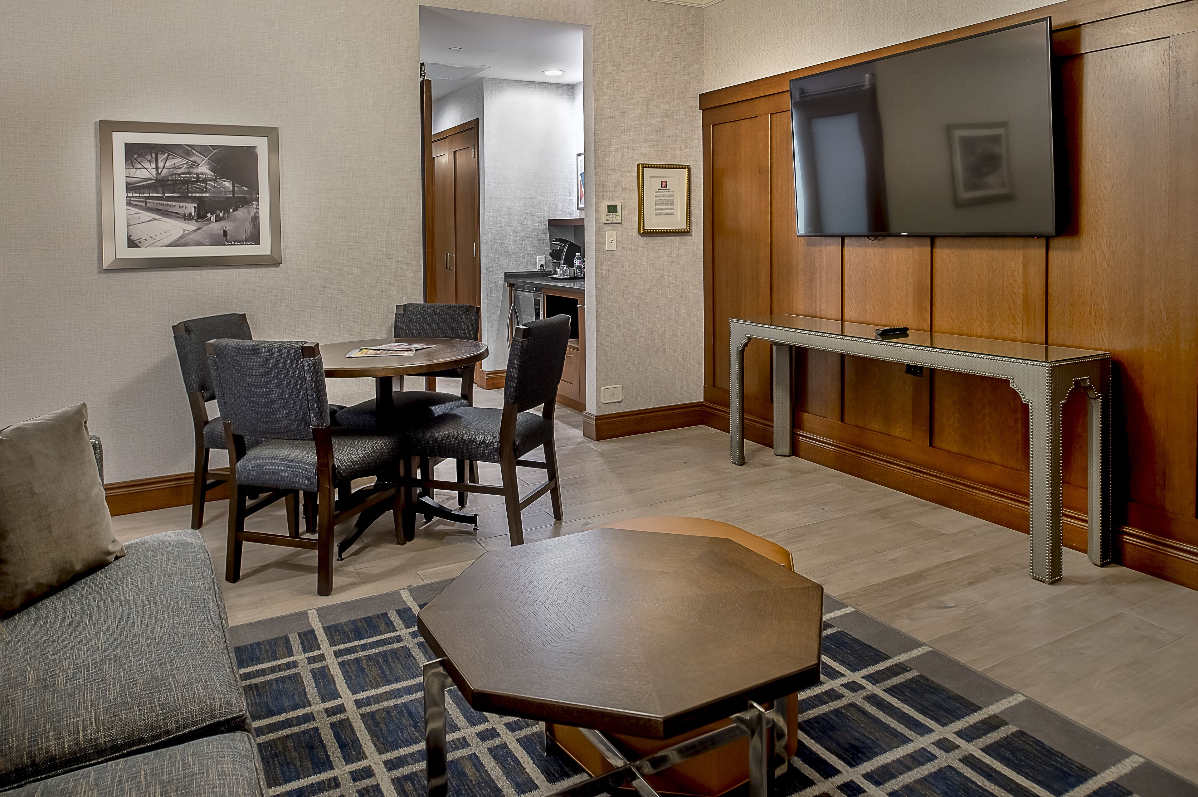 A hotel room with a dining area, a sofa, a TV mounted on the wall, and a framed picture on the wall.