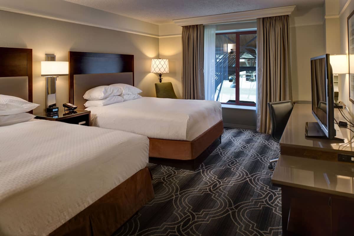 A hotel room with two beds, a desk, a chair, a TV, and a window with curtains.