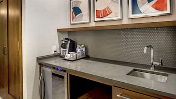 Una cocina con un lavamanos de acero inoxidable, una cafetera y un lavavajillas. Hay obras de arte enmarcadas en la pared sobre la encimera.