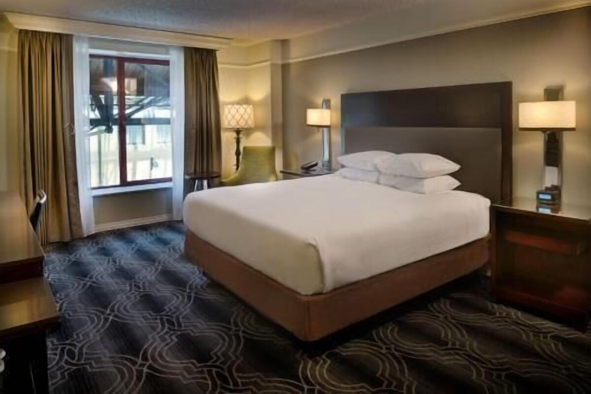 A hotel room with a large bed, two bedside lamps, a nightstand, a window with curtains, and a patterned carpet.