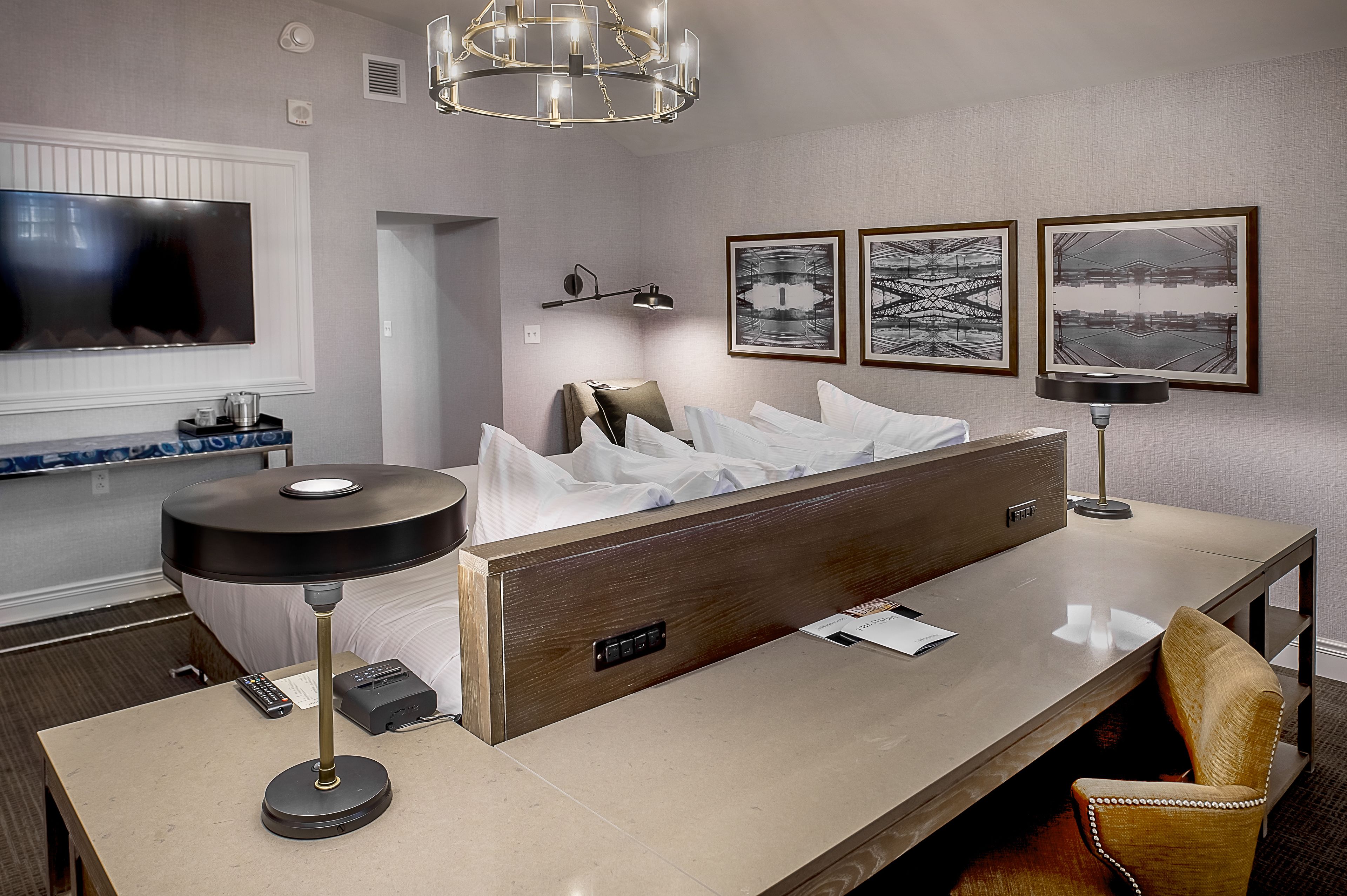 A modern hotel room with a large bed, a desk with a lamp, a television, and artwork on the wall.