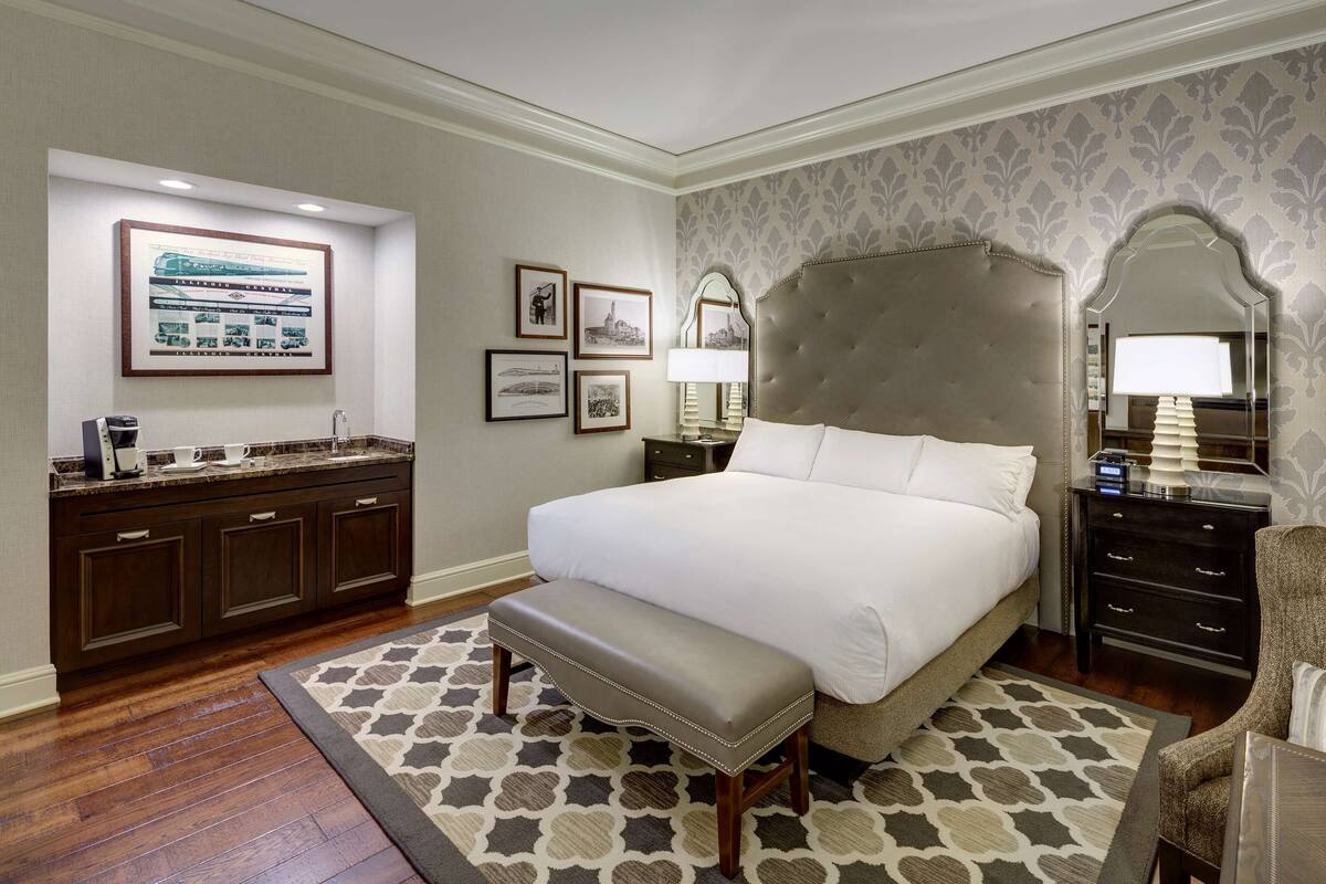 A hotel room with a large bed, a bench, a dresser, and a mirror.