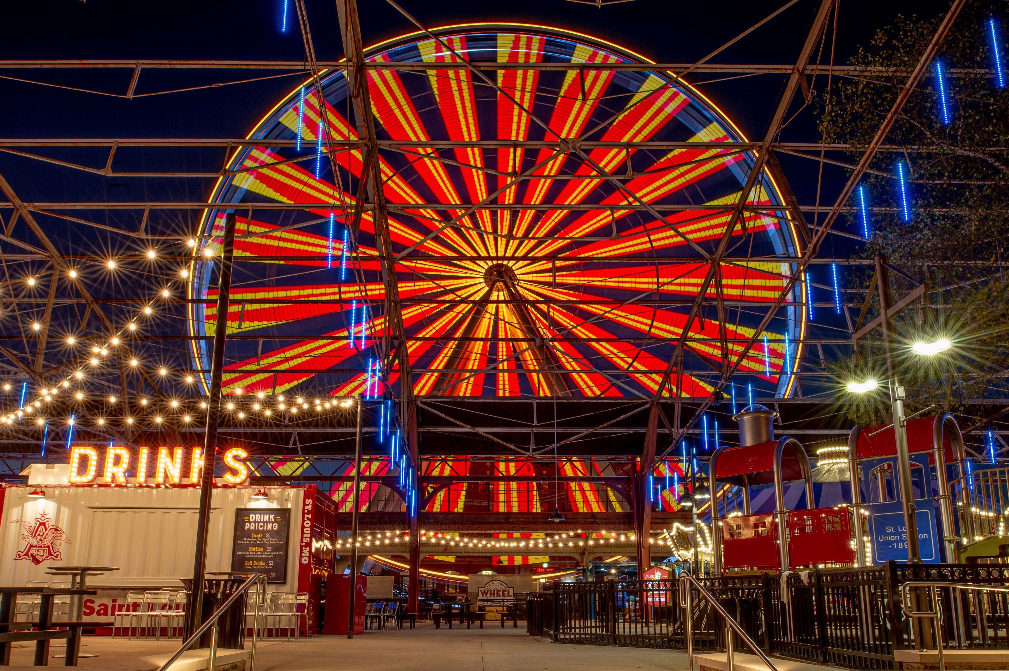 A large Ferris wheel with a colorful light display, surrounded by a covered area with a 'Drinks' sign and a 'Saint Louis' sign.