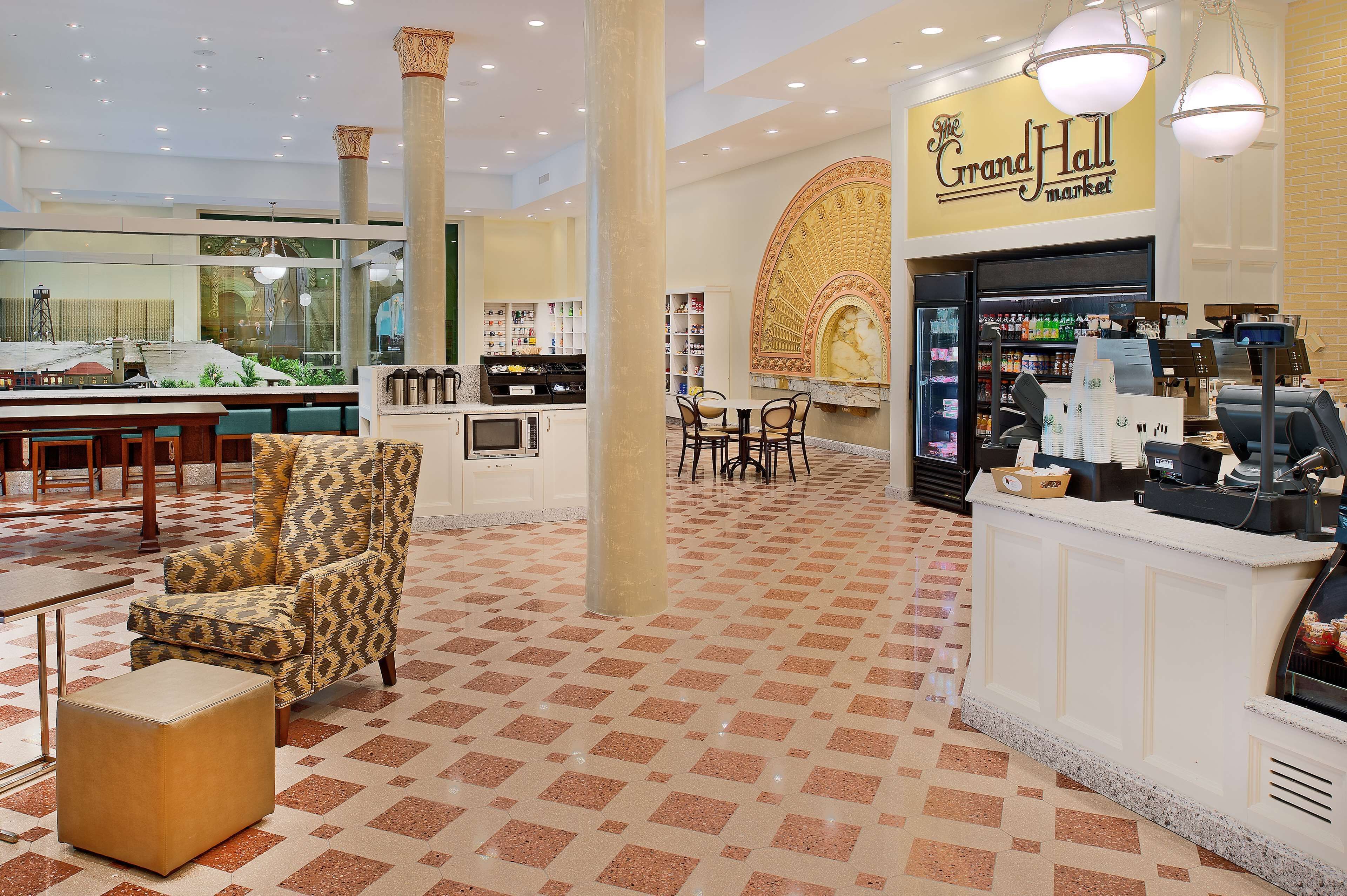 Een ruime lobby met een zwart-wit schachoot patroon, een bord met de tekst Grand Hall Market, een zithoek met gestreepte stoelen en een koffiehoek.