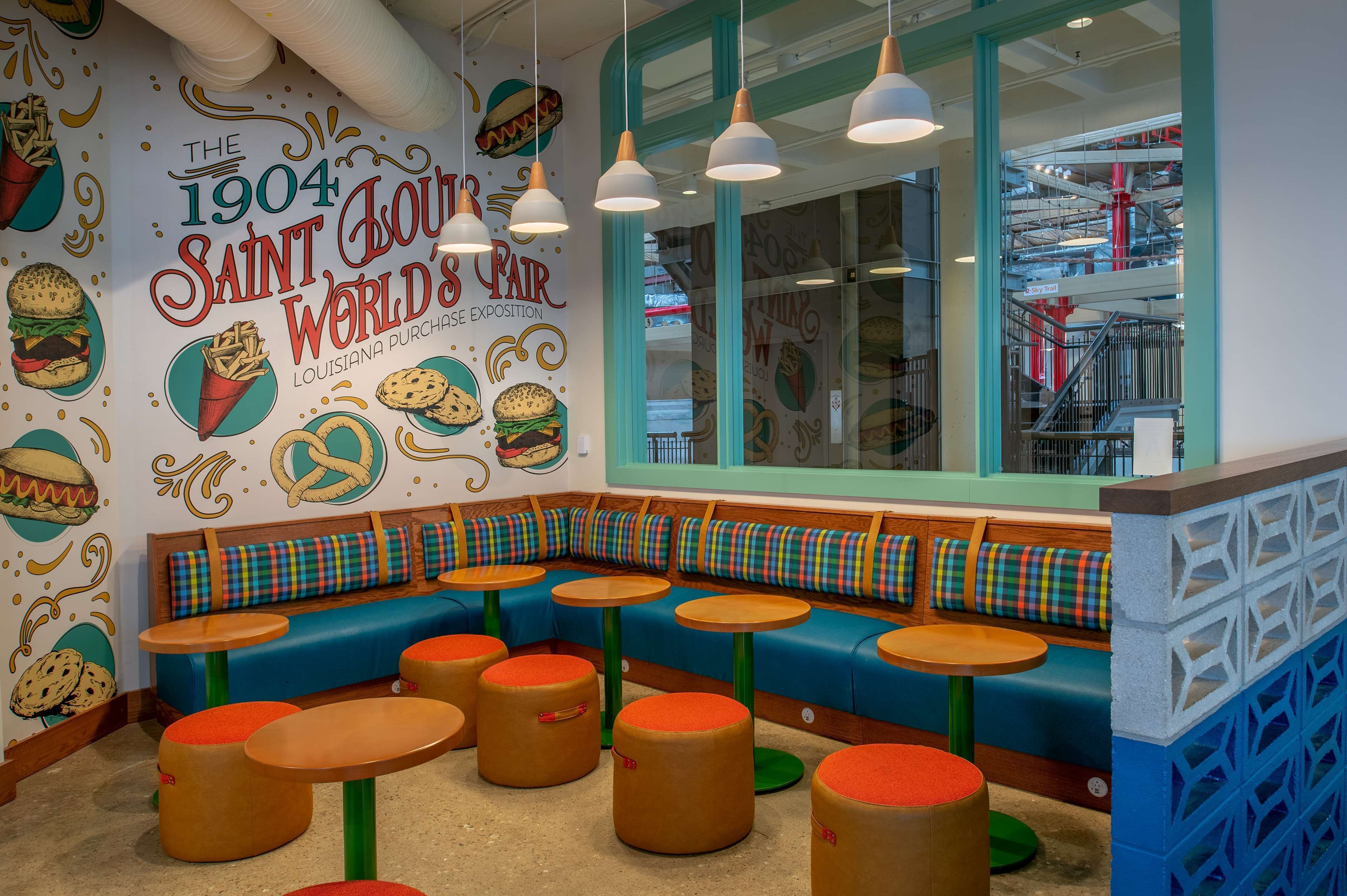 Interior de un restaurante con un mural de pared colorido, una zona de estar con bancos y mesas redondas, y vista del interior del edificio a través de una ventana.