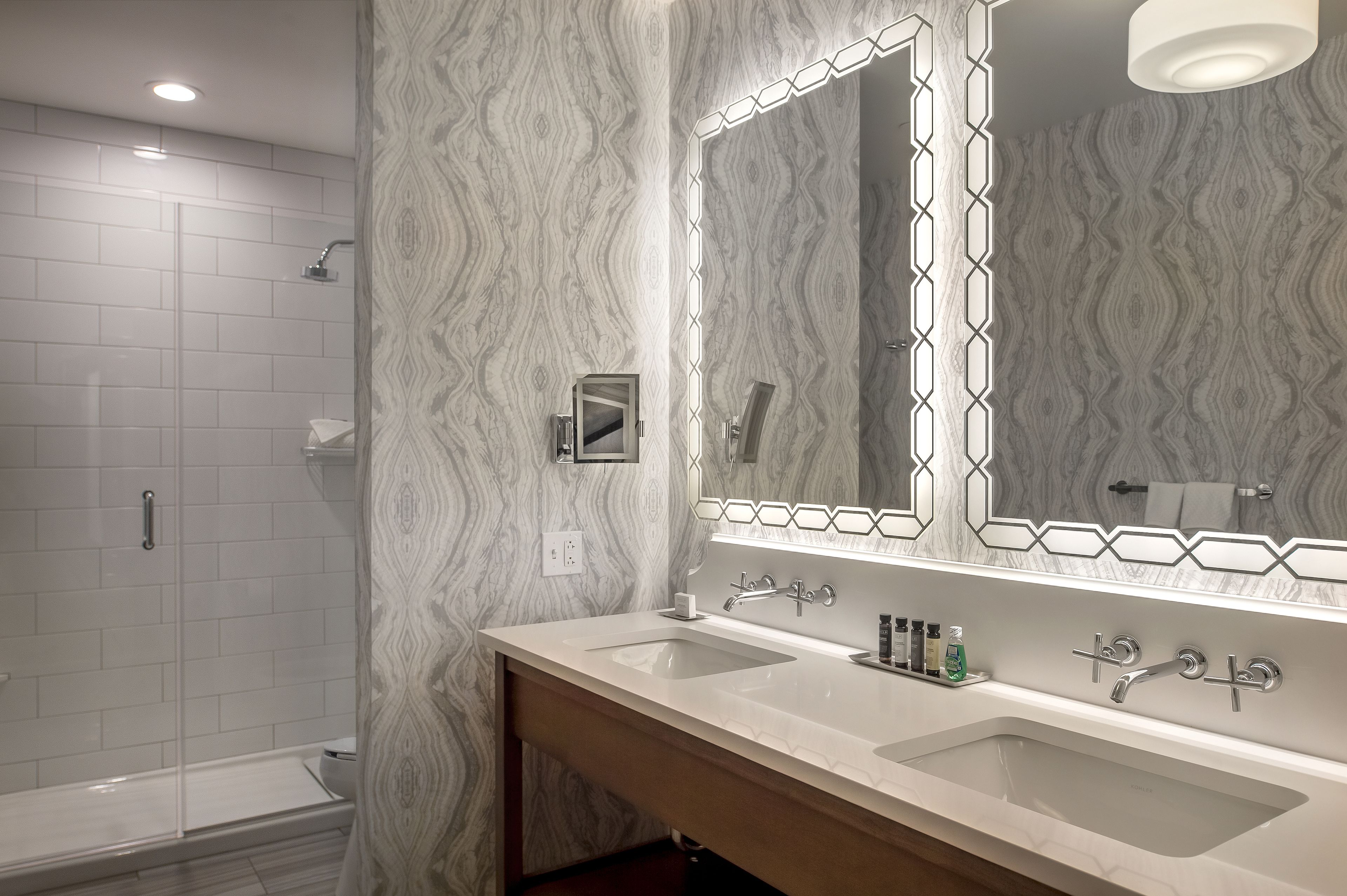 Un bagno con doppio lavandino, uno specchio grande illuminato, una doccia e carta da parati decorata.