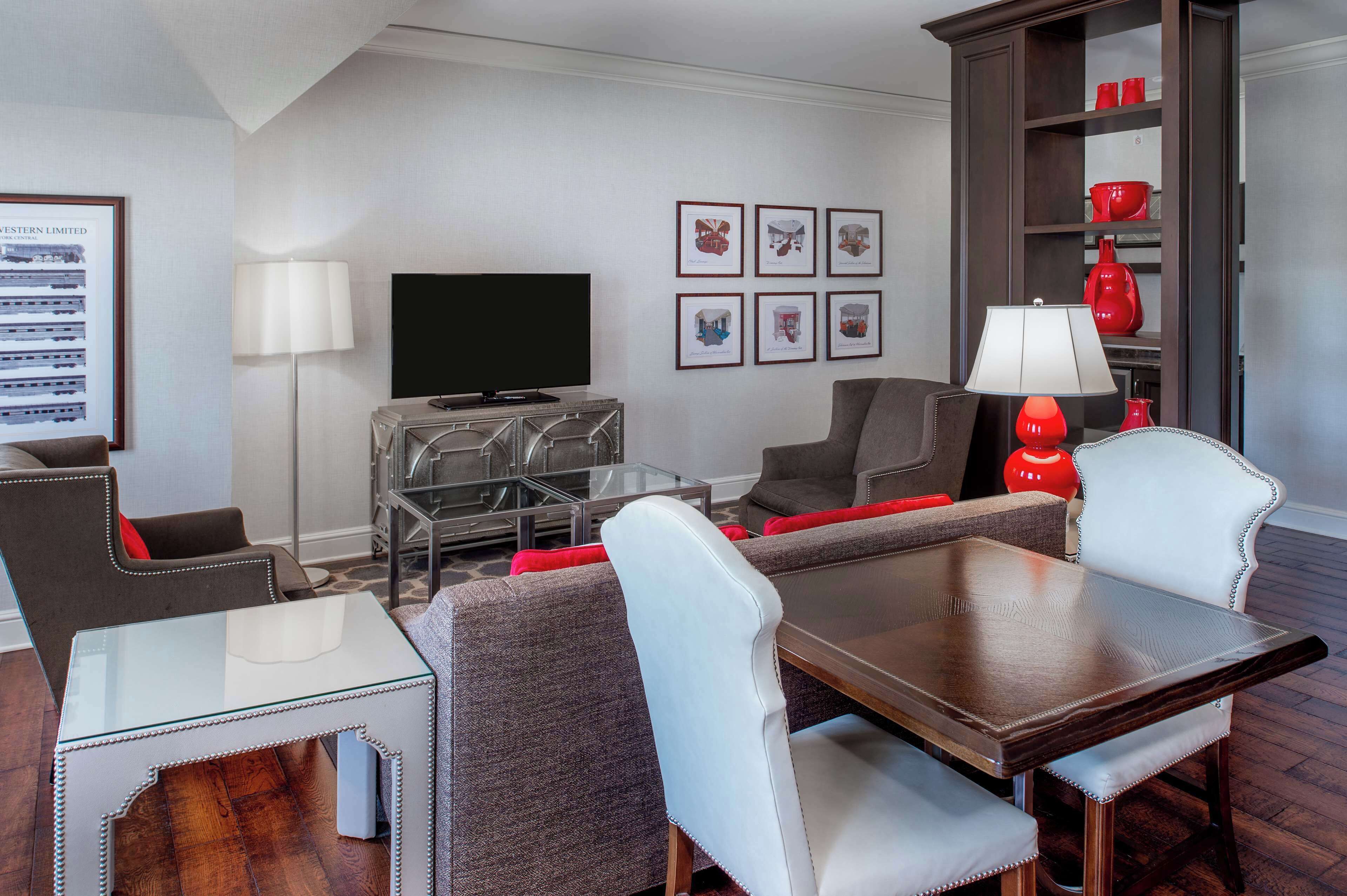 A modern living room with a glass coffee table, grey and white upholstered chairs, a wooden dining table, a red lamp, a flat-screen TV, and a bookshelf with red decorative items.
