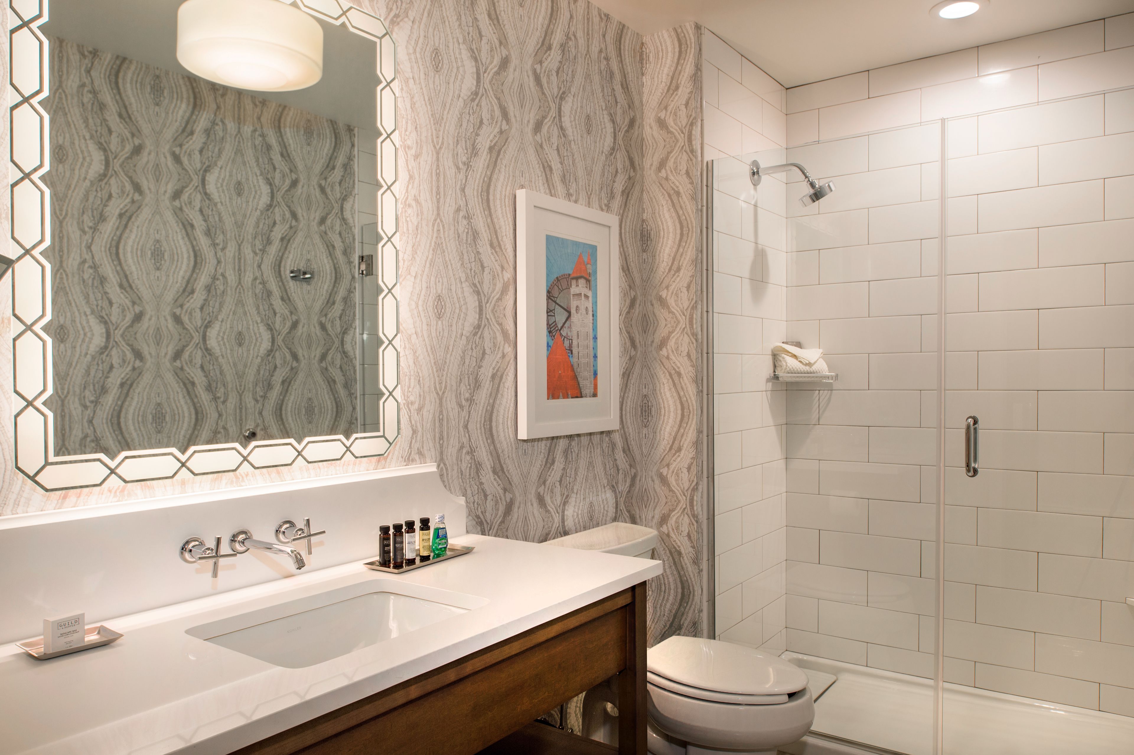 A bathroom with a marble-patterned wall, a large mirror, a sink with a vanity, a toilet, and a glass-enclosed shower.