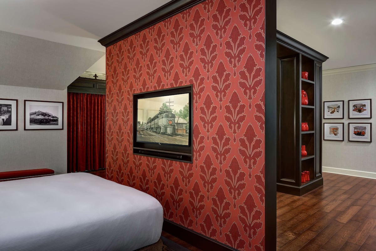 A hotel room with a bed, a television mounted on the wall, a red patterned wallpaper, and a wooden floor.