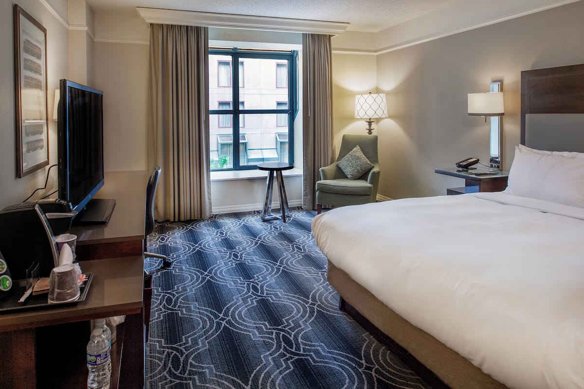 A hotel room with a large bed, a desk with a television and coffee maker, a chair, a nightstand with a lamp, and a window with curtains.