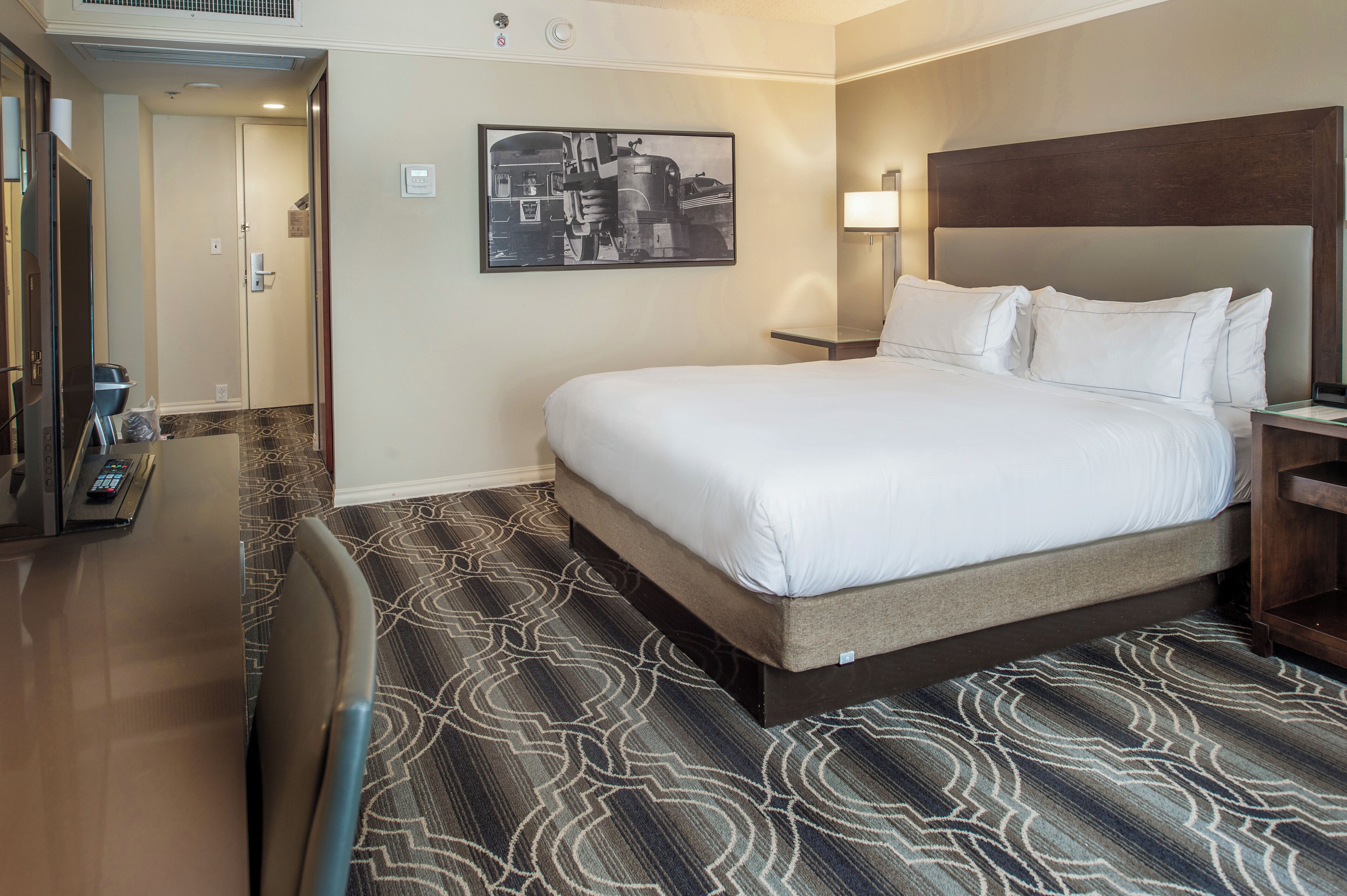 Quarto de hotel com uma cama grande, uma escrivaninha com cadeira, um quadro na parede e um tapete estampado.