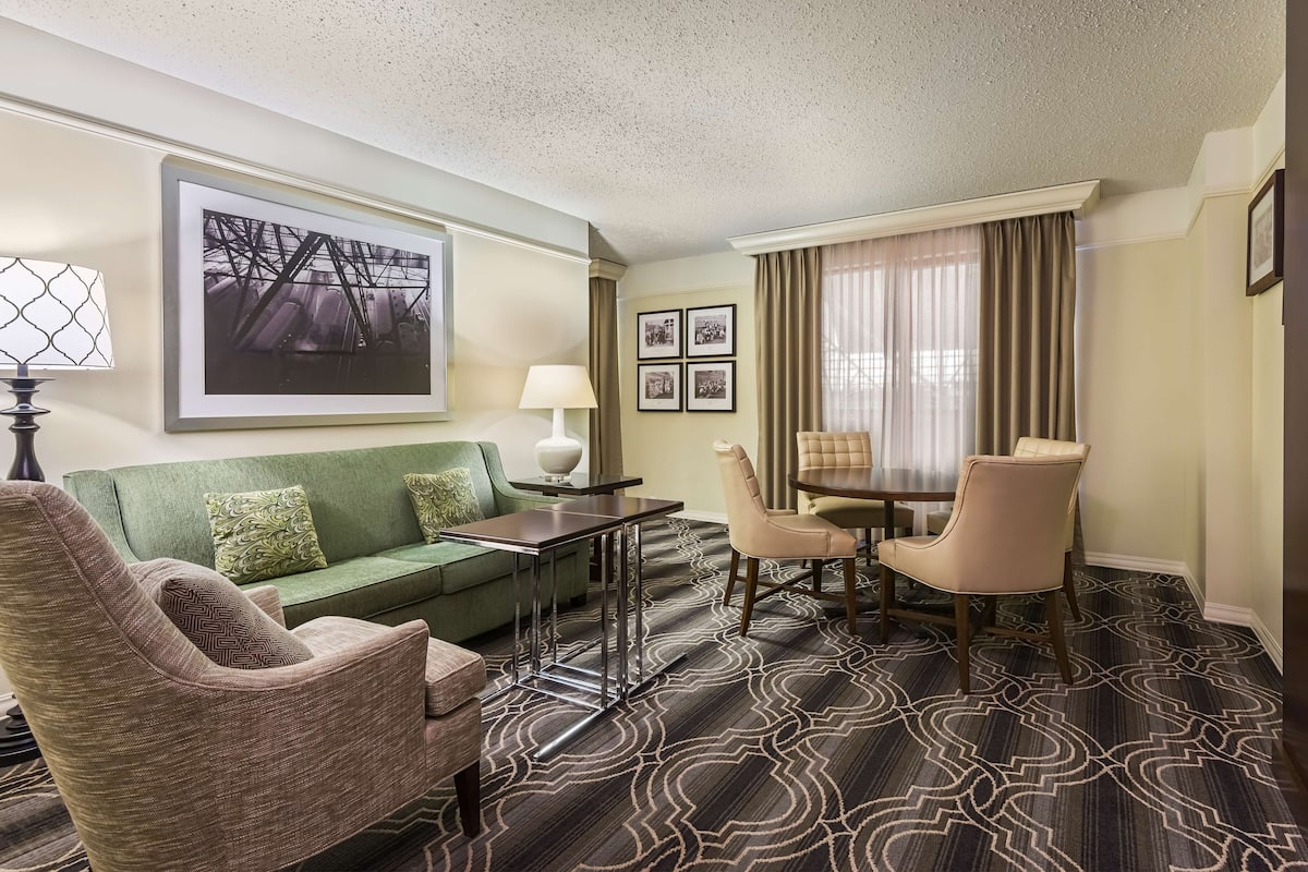 A hotel room with a green sofa, a beige armchair, a dining table with chairs, a lamp, and a large framed picture on the wall.