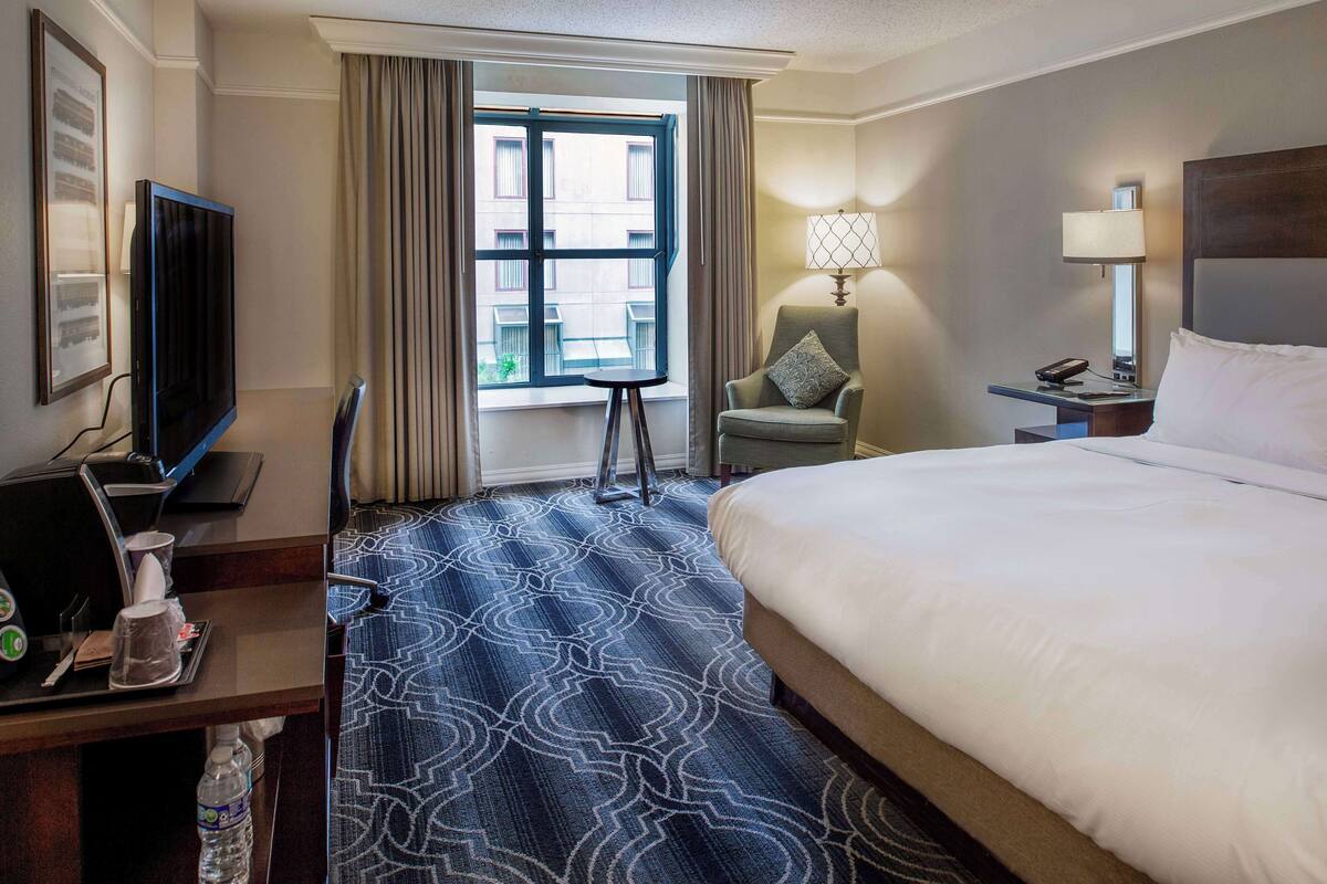 A hotel room with a large bed, a desk with a television, a chair, a small table, a lamp, and a window with curtains.