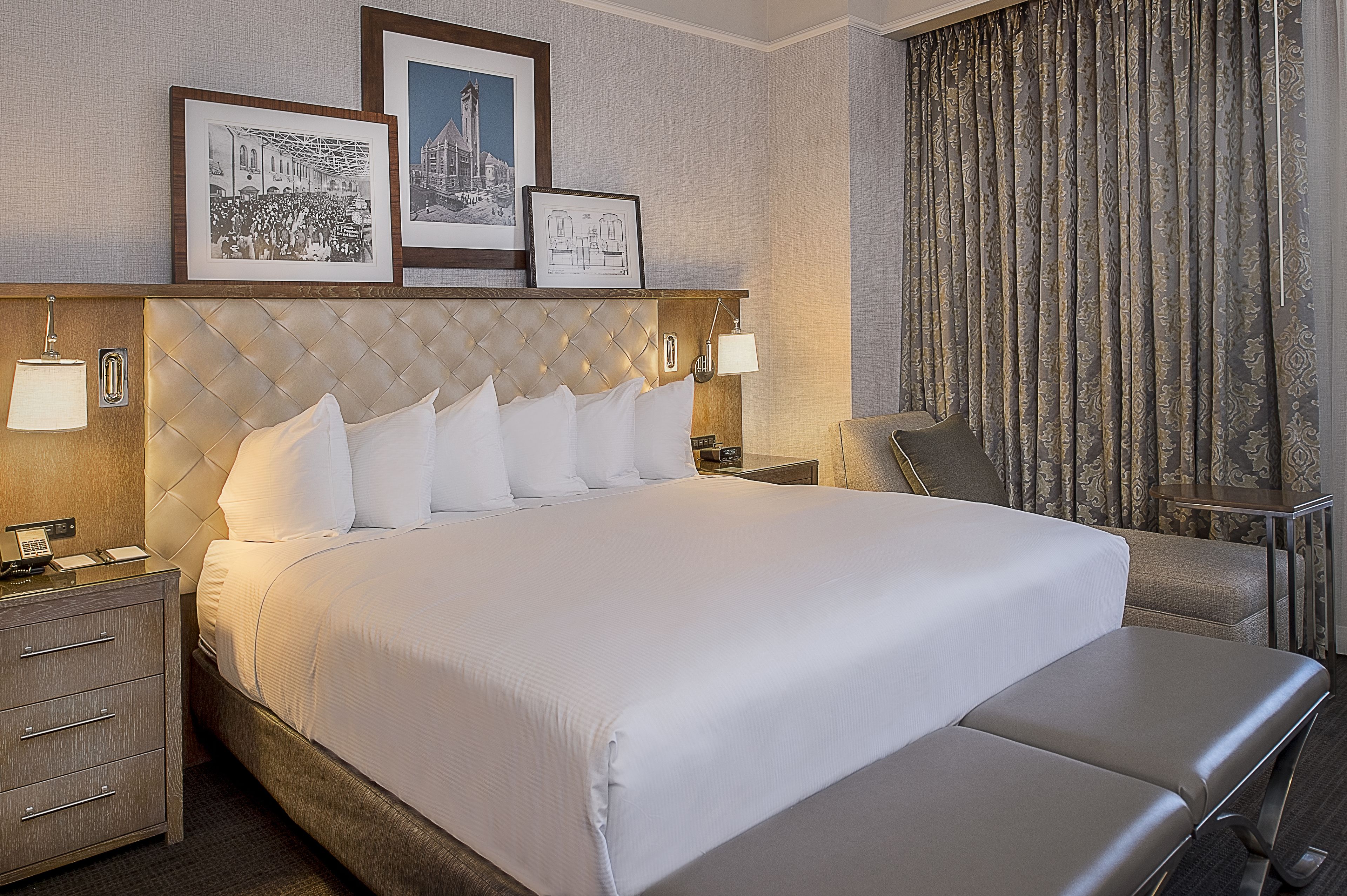 A neatly made bed with white linens, a tufted headboard, and a bedside table with a lamp. There are framed pictures above the bed and a curtain on the window.