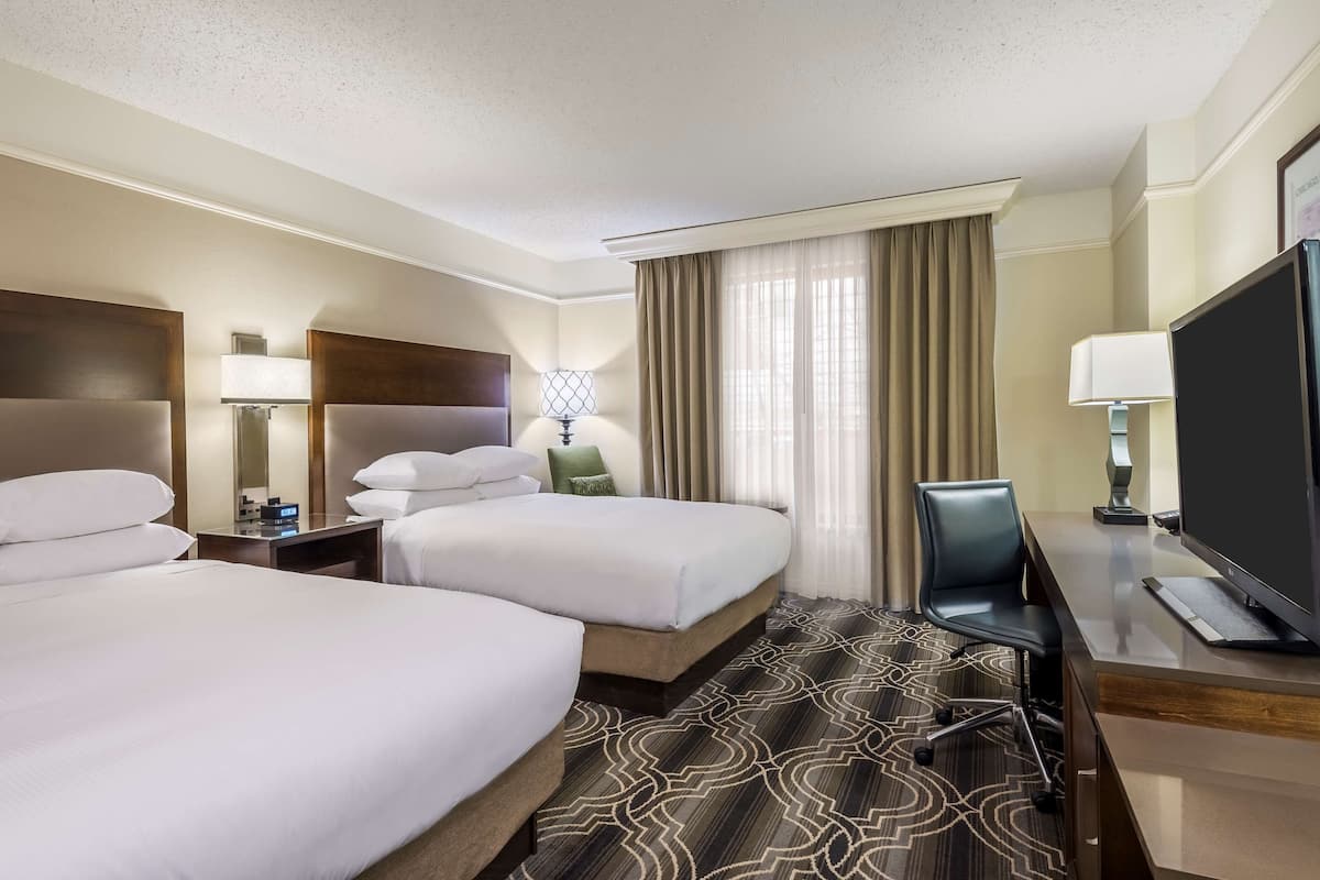 A hotel room with two beds, a desk with a TV, a chair, a lamp, and a window with curtains.