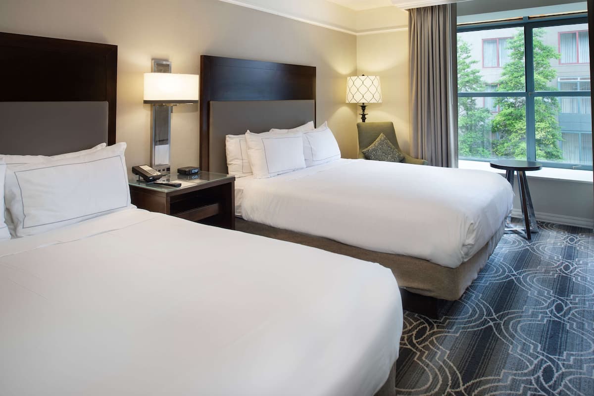 A hotel room with two beds, a nightstand, a lamp, and a view of the outside.