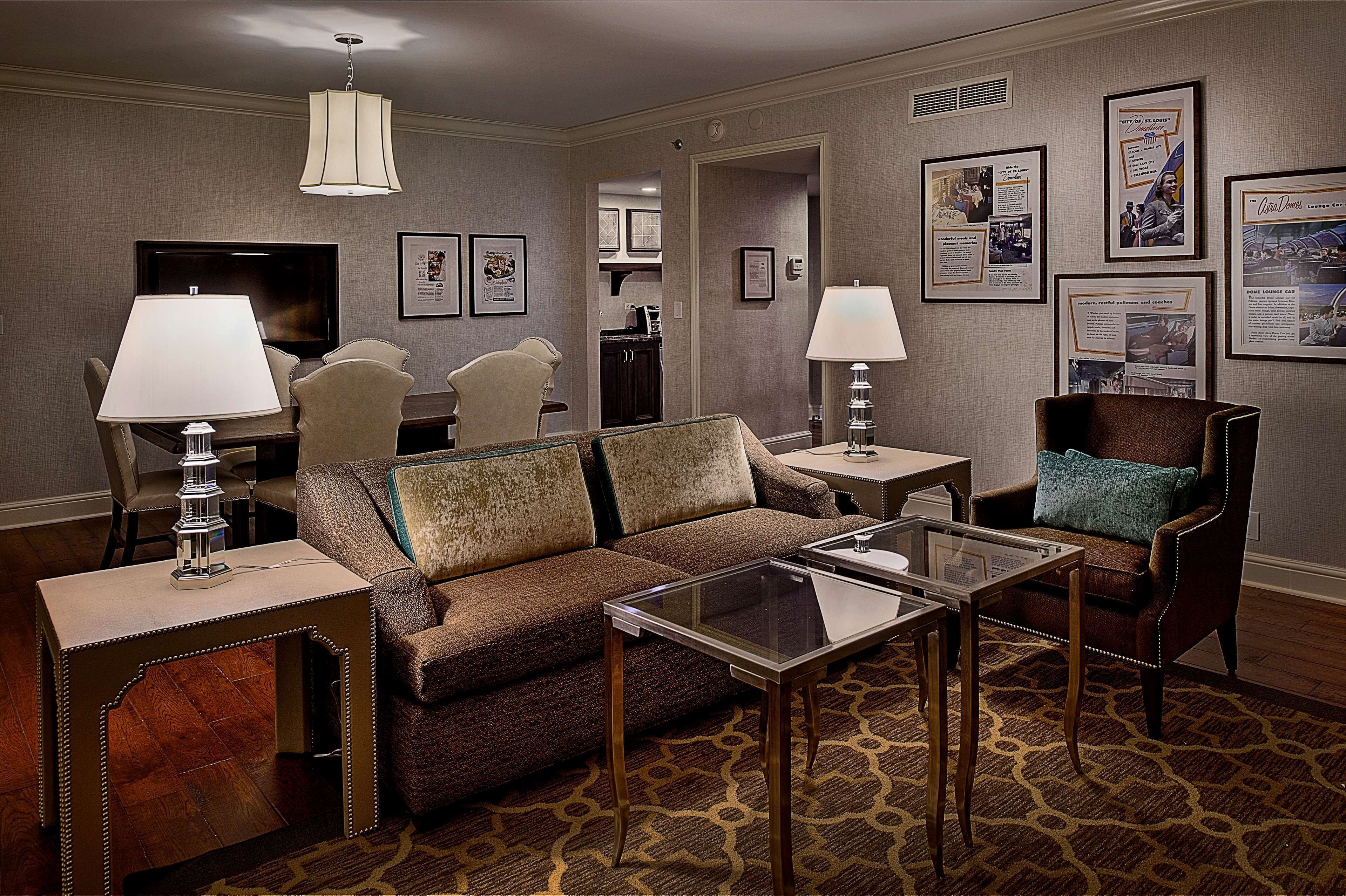 A hotel room with a brown sofa, two armchairs, a glass coffee table, and a TV. There are framed pictures on the walls and a lamp on each side of the sofa.