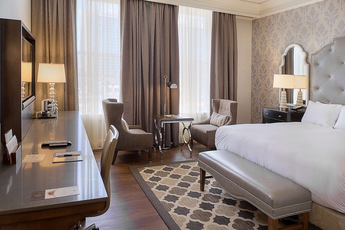 A hotel room with a large bed, a desk with a lamp, a chair, a small table, and a bench.