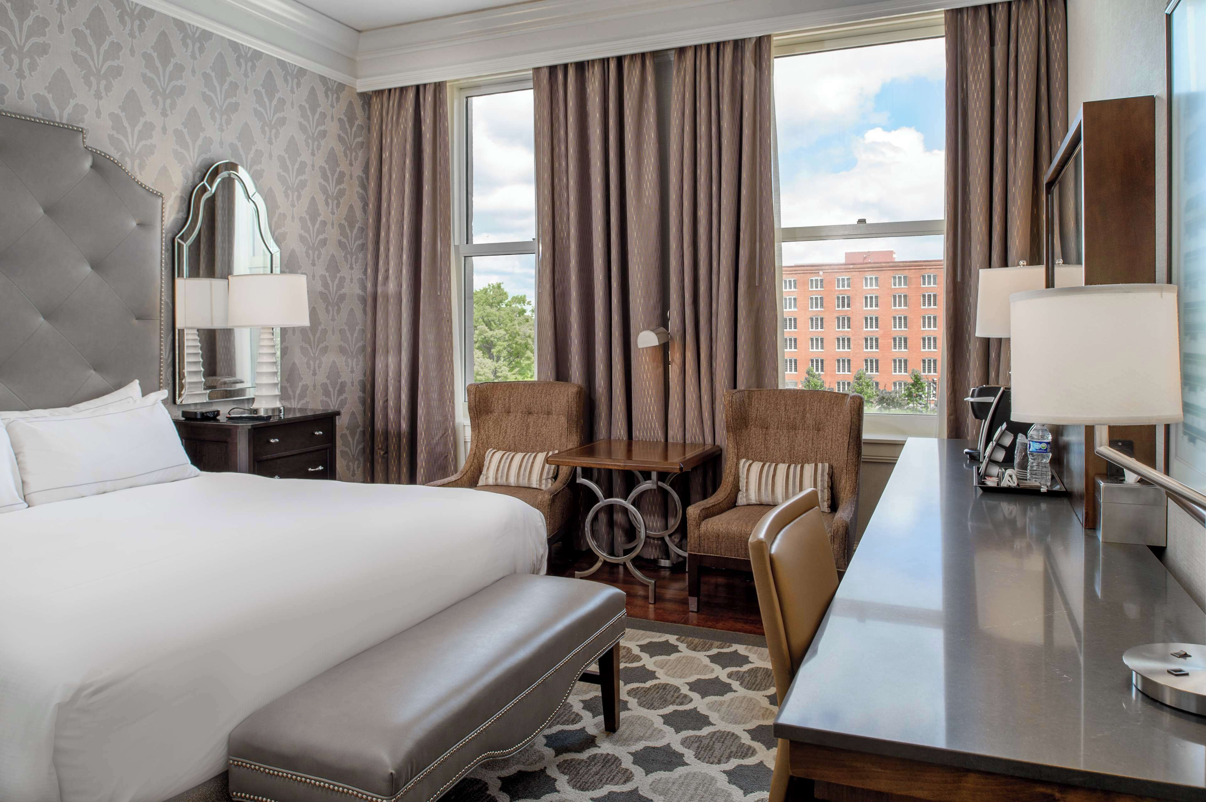 Habitación de hotel con una cama grande, un escritorio, dos sillones, un espejo y vistas a un edificio a través de la ventana.