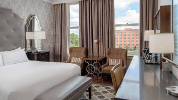Habitación de hotel con una cama grande, un escritorio, dos sillones, un espejo y vistas a un edificio a través de la ventana.