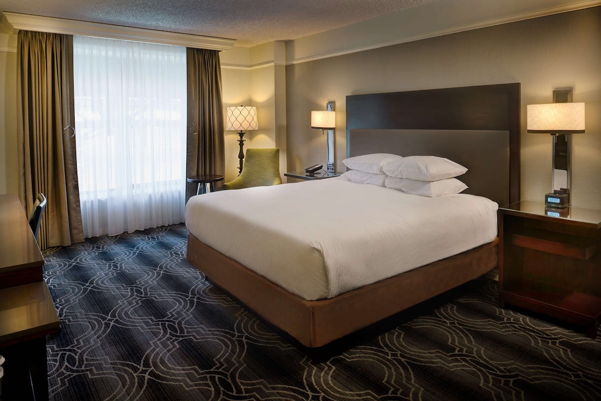 A hotel room with a large bed, two bedside tables with lamps, a chair, a desk, and a large window with curtains.