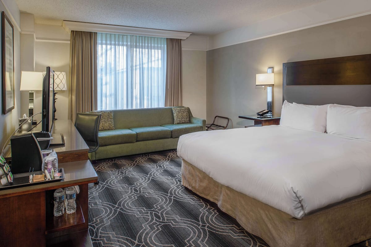A hotel room with a large bed, a sofa, a desk with a computer, a lamp, and a window with curtains.