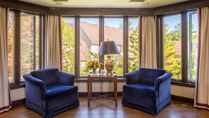 A room with two blue armchairs, a small table with a lamp and vase, and large windows with curtains. - The Orchards Hotel (Williamstown)
